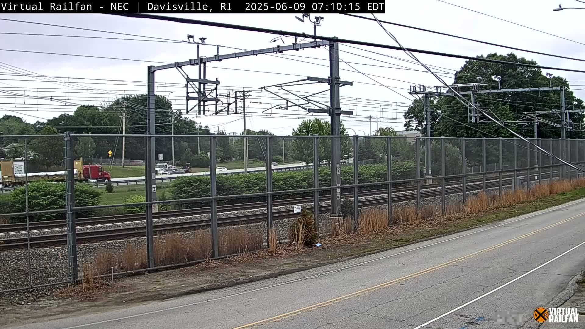 A mostly cloudy day shows a view of railroad tracks behind a chain-link fence, with a road in the foreground and several vehicles visible in the distance.