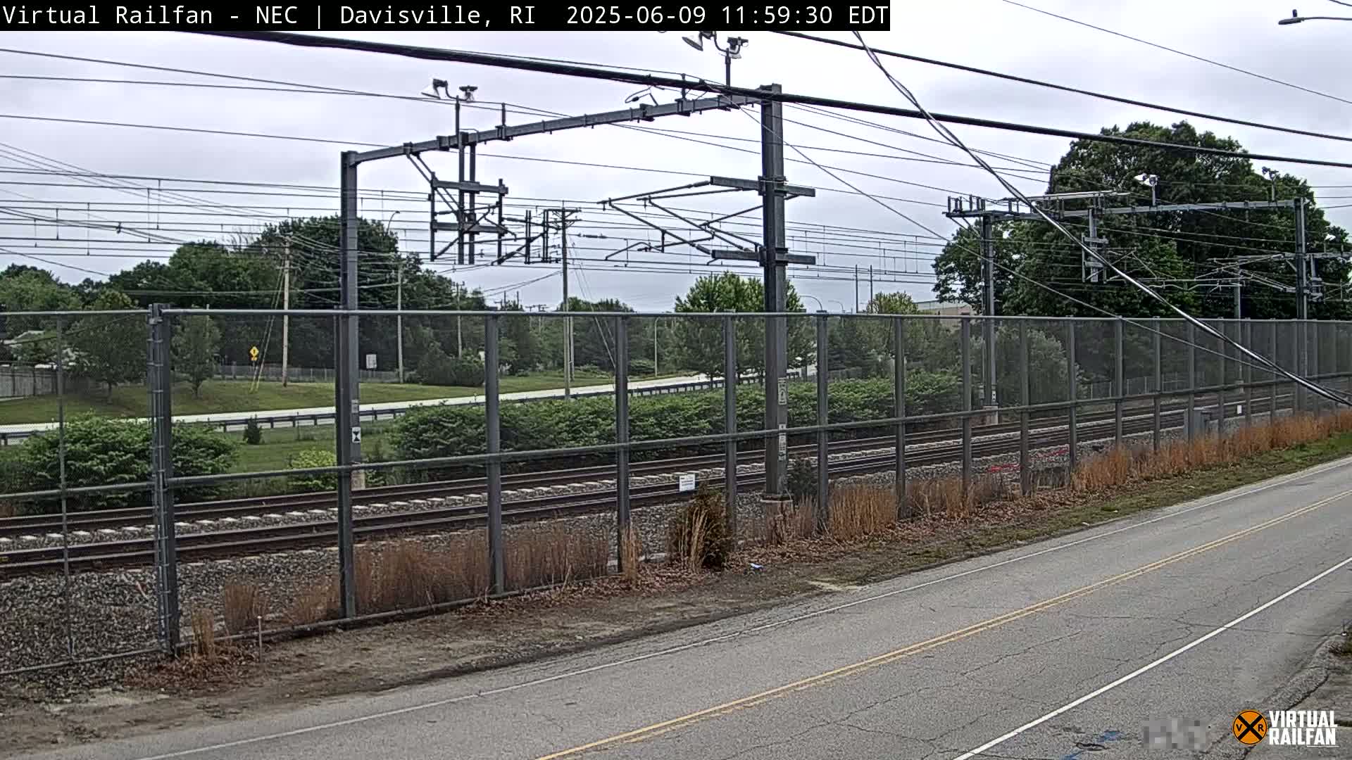 A view of railroad tracks behind a chain-link fence on an overcast day, with a road in the foreground.