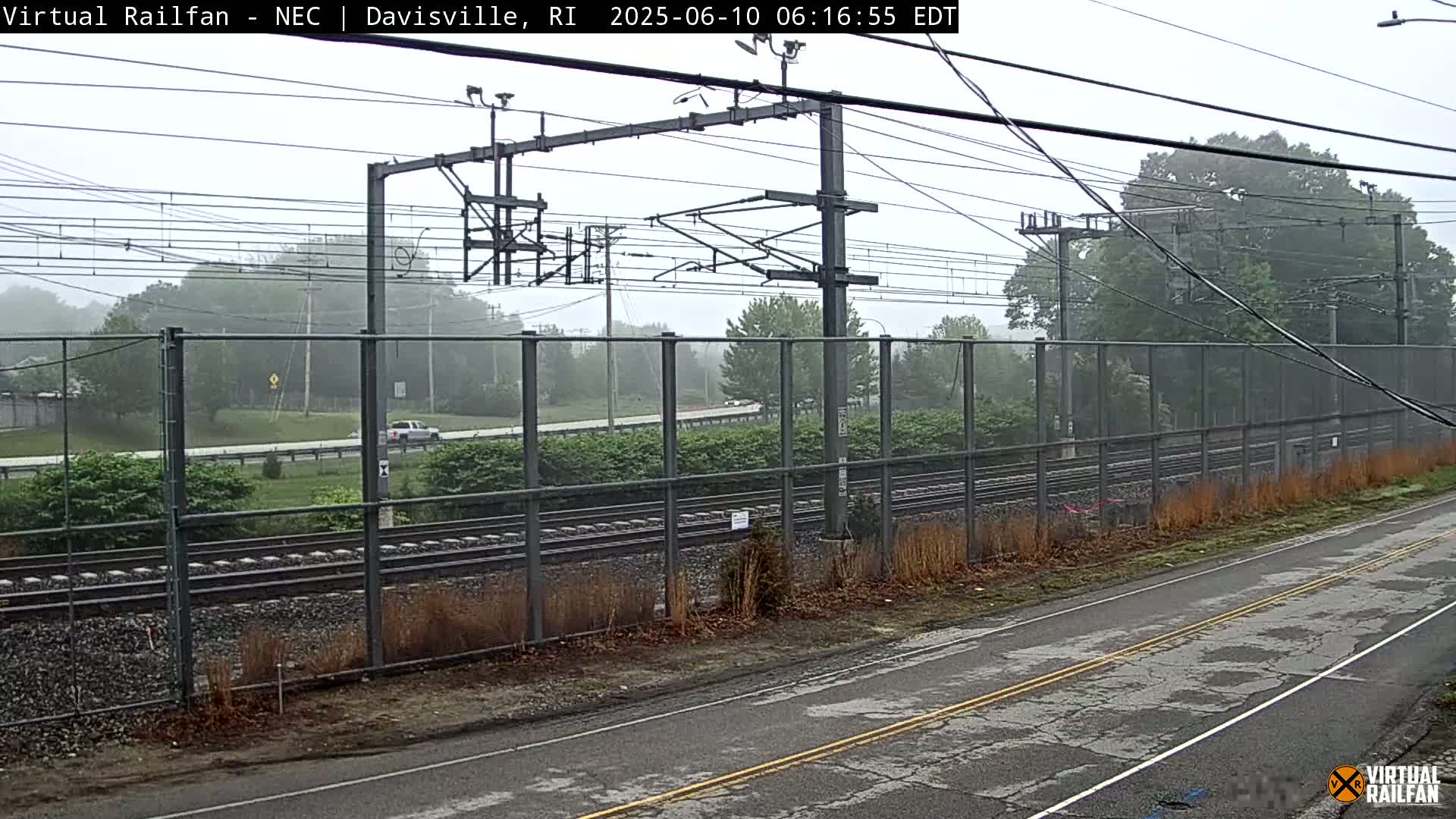 A view of train tracks behind a chain-link fence on an overcast day, with a road in the foreground and a vehicle visible in the distance.