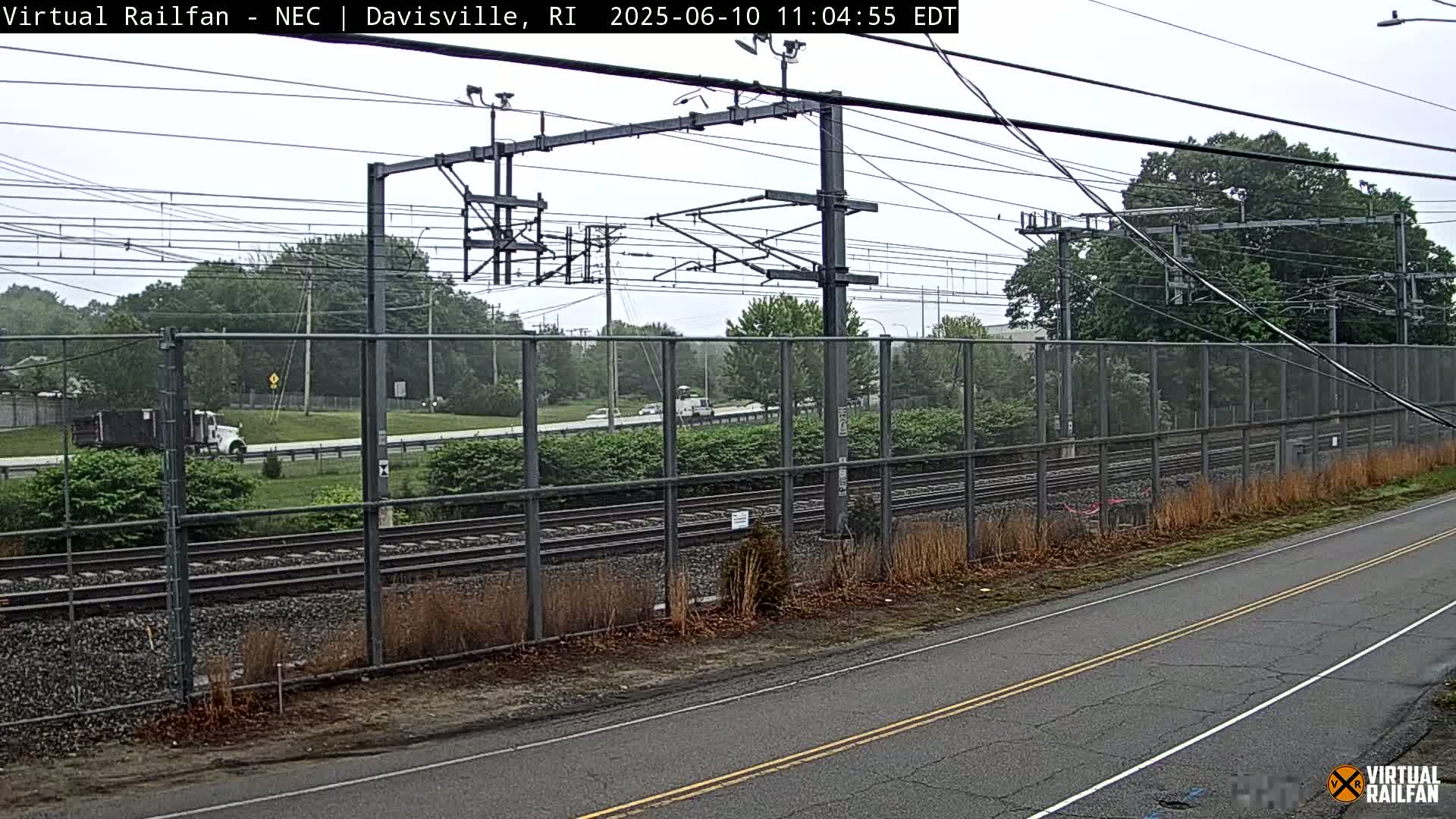 A view of train tracks and a road beyond a chain-link fence on an overcast day.