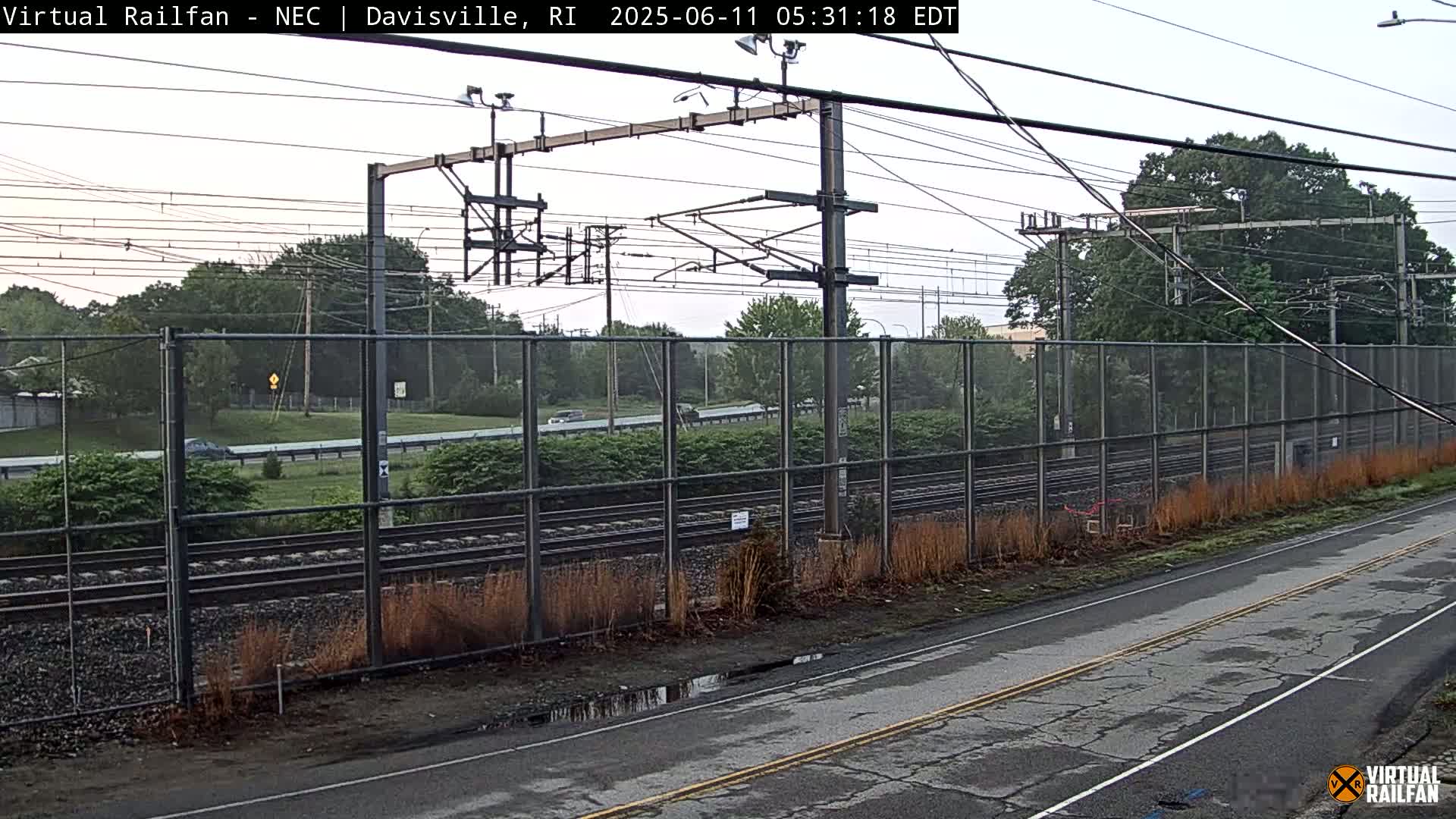 A view of railroad tracks behind a chain-link fence on an overcast day, with a road in the foreground.