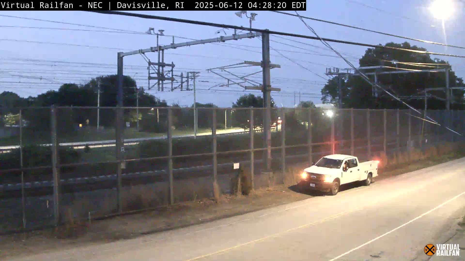 A white pickup truck is parked on a road next to a chain-link fence, beyond which are train tracks and power lines under a twilight sky.