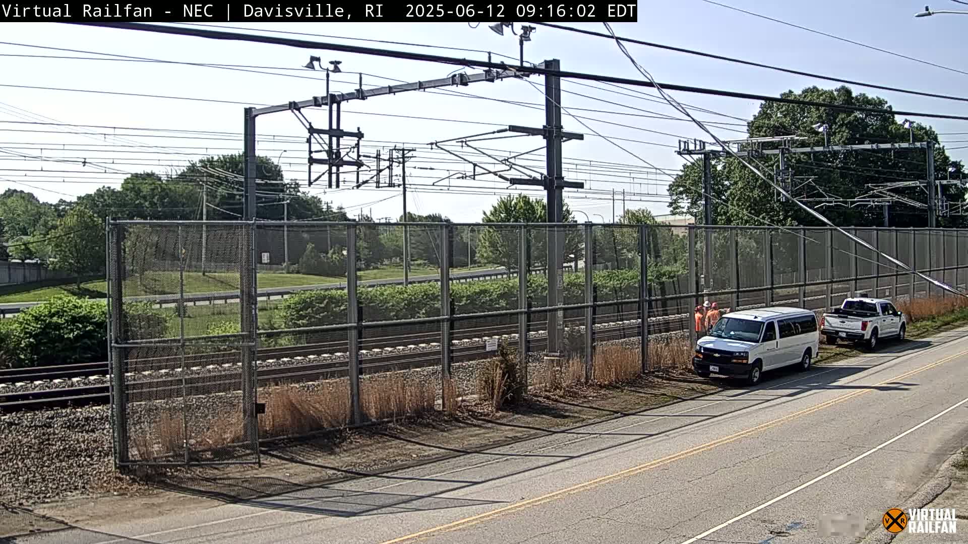 A sunny day reveals railroad tracks beside a road, fenced in with a chain link fence, and several workers near a white van and a pickup truck.