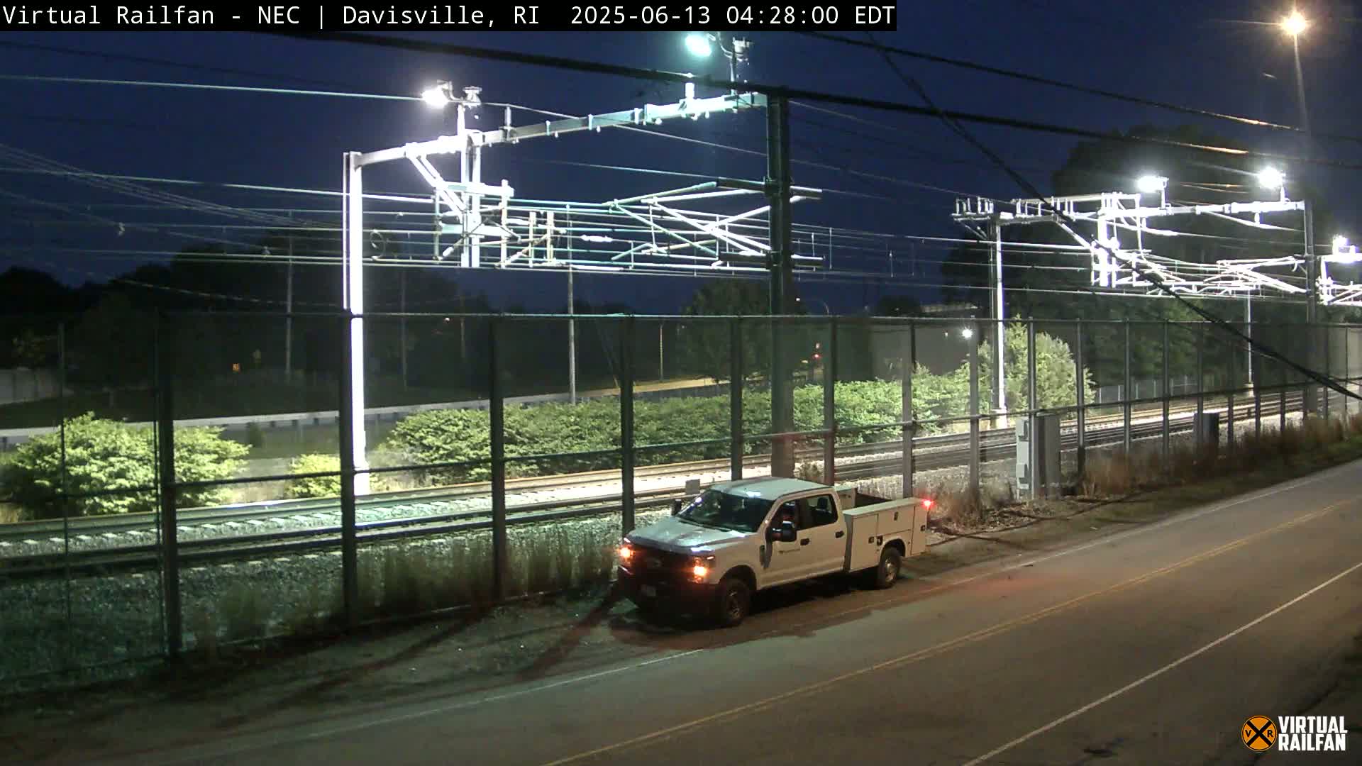 A white pickup truck is parked near a fenced-in railway at night, under bright lights.