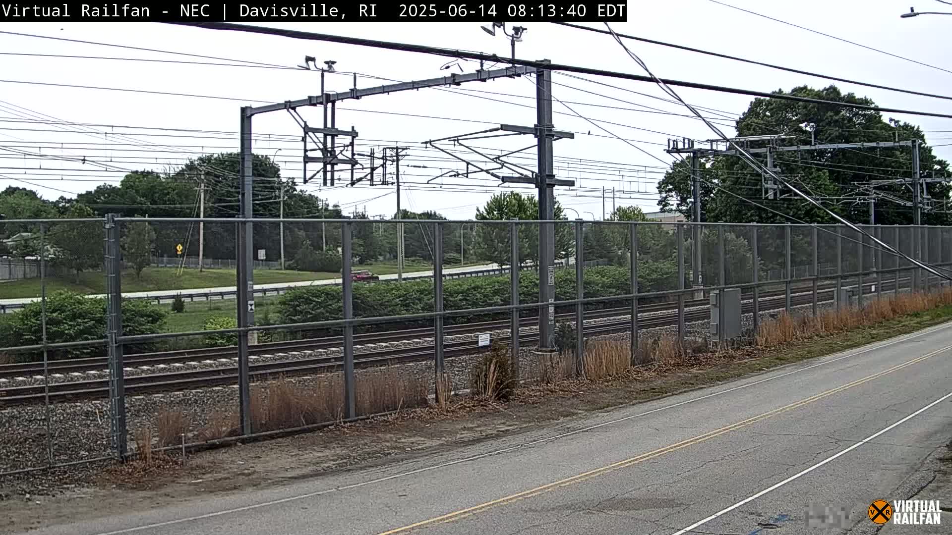 A view of railroad tracks behind a chain-link fence on an overcast day, with a road in the foreground.