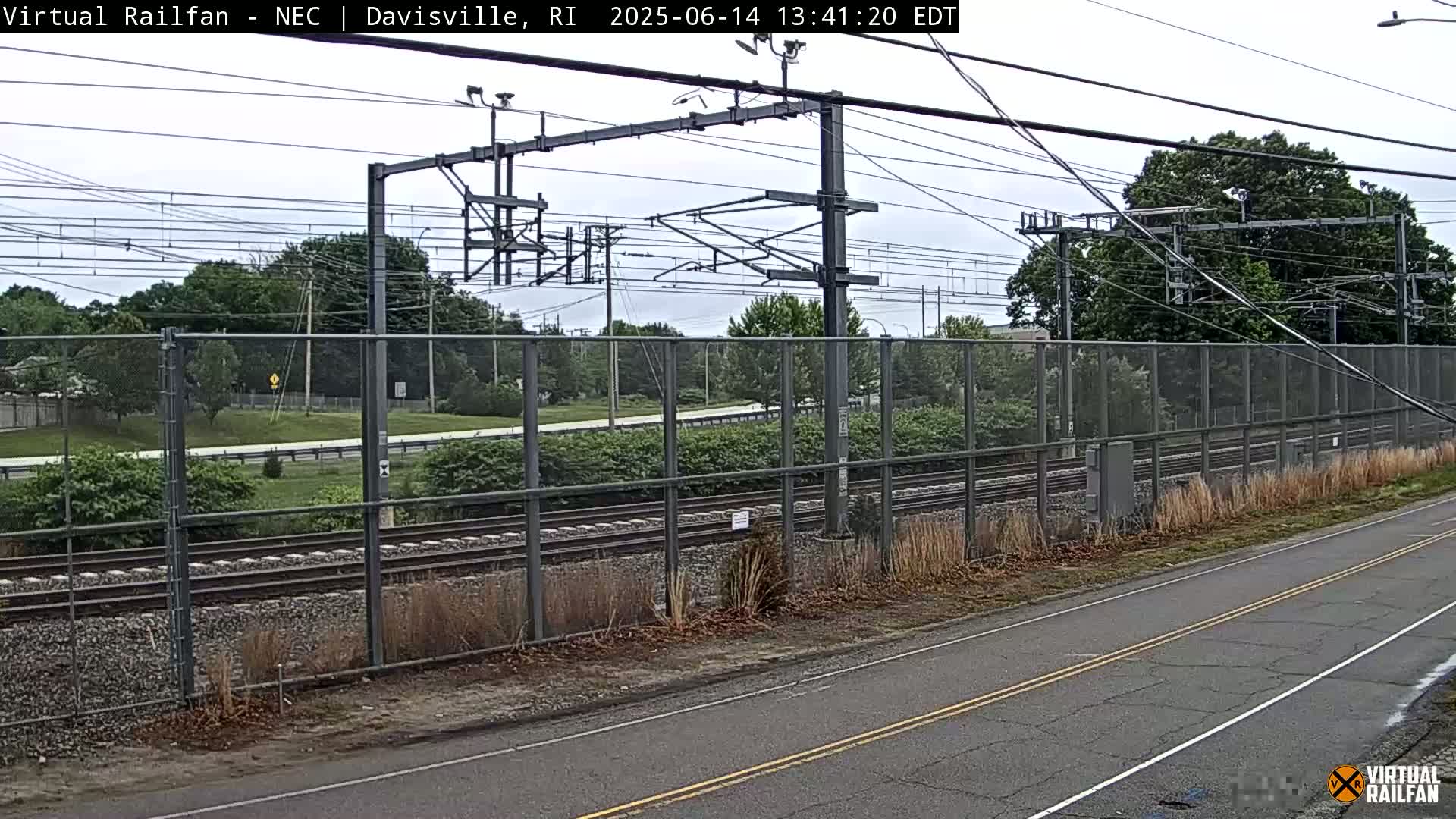 A view of a railroad track behind a chain link fence on an overcast day, with a road in the foreground.