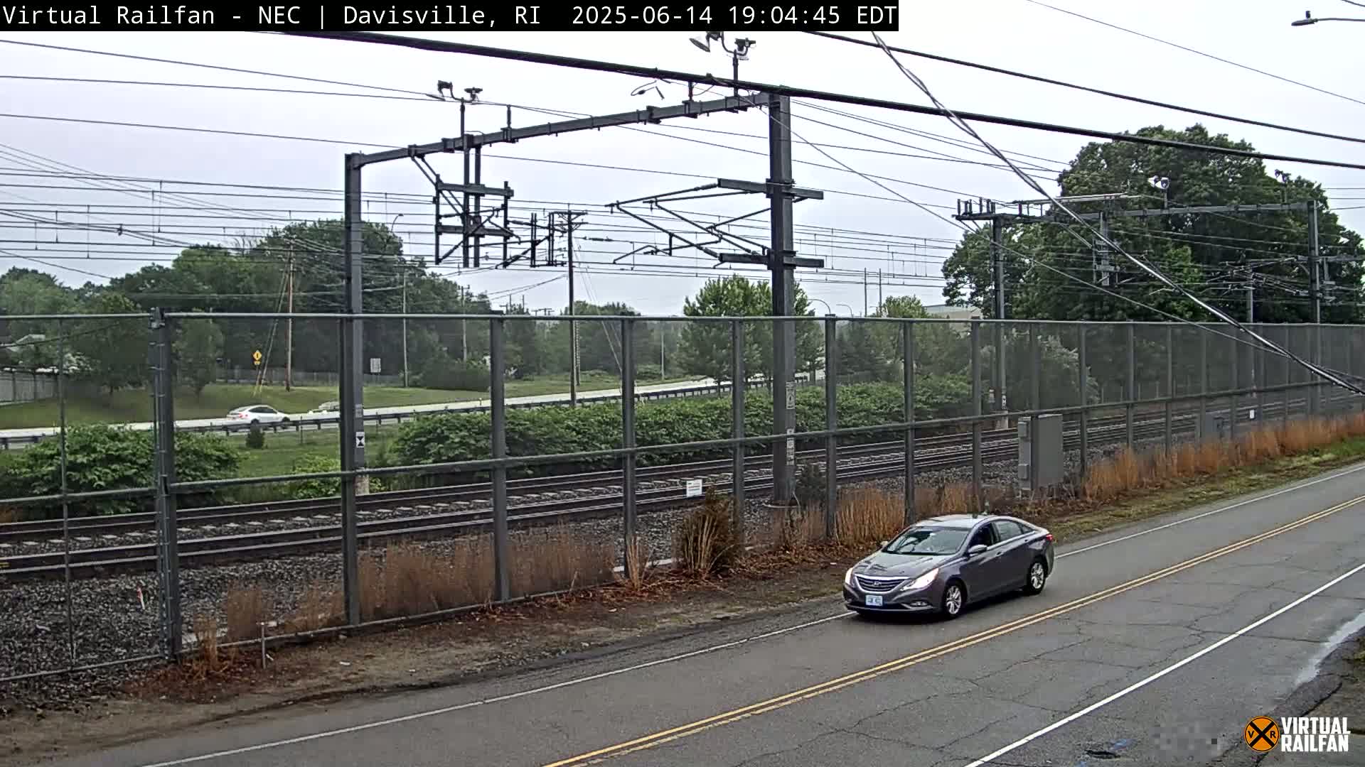 A view of a railroad track behind a chain link fence on an overcast day, with a road in the foreground.