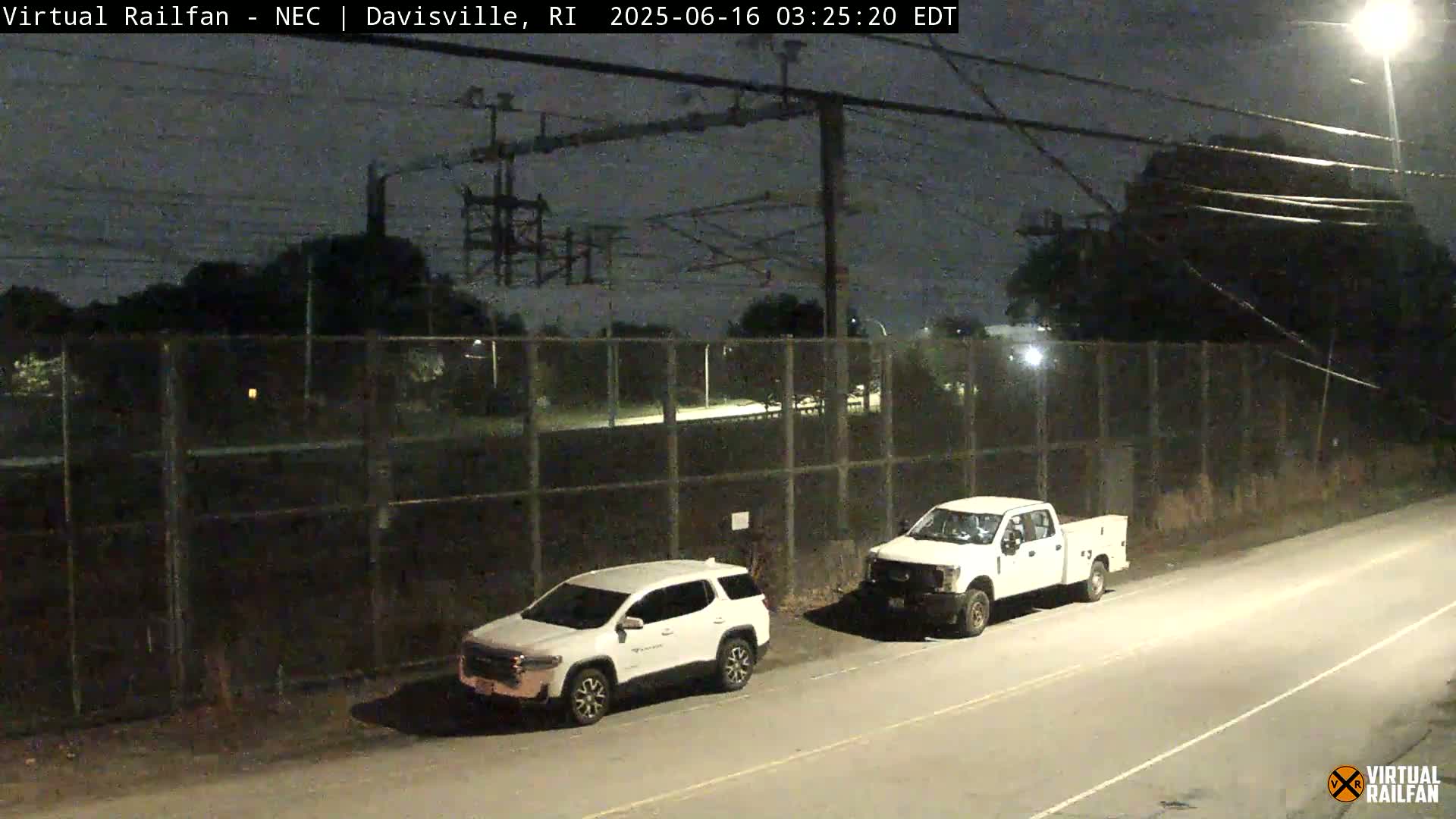 At night, two white vehicles are parked near a chain-link fence beside a road under a dark sky.