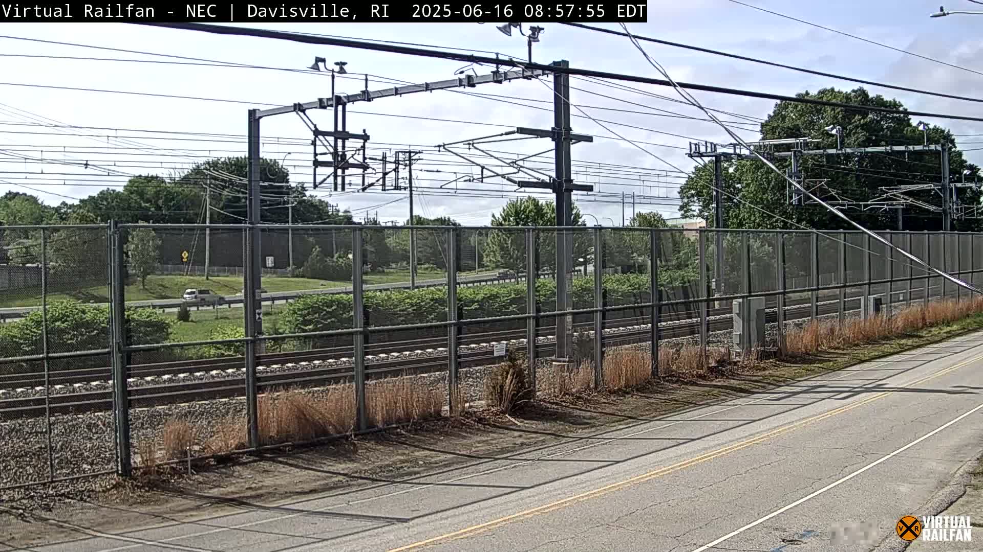 A view of a roadway beside a chain-link fence enclosing railroad tracks under a partly cloudy sky.