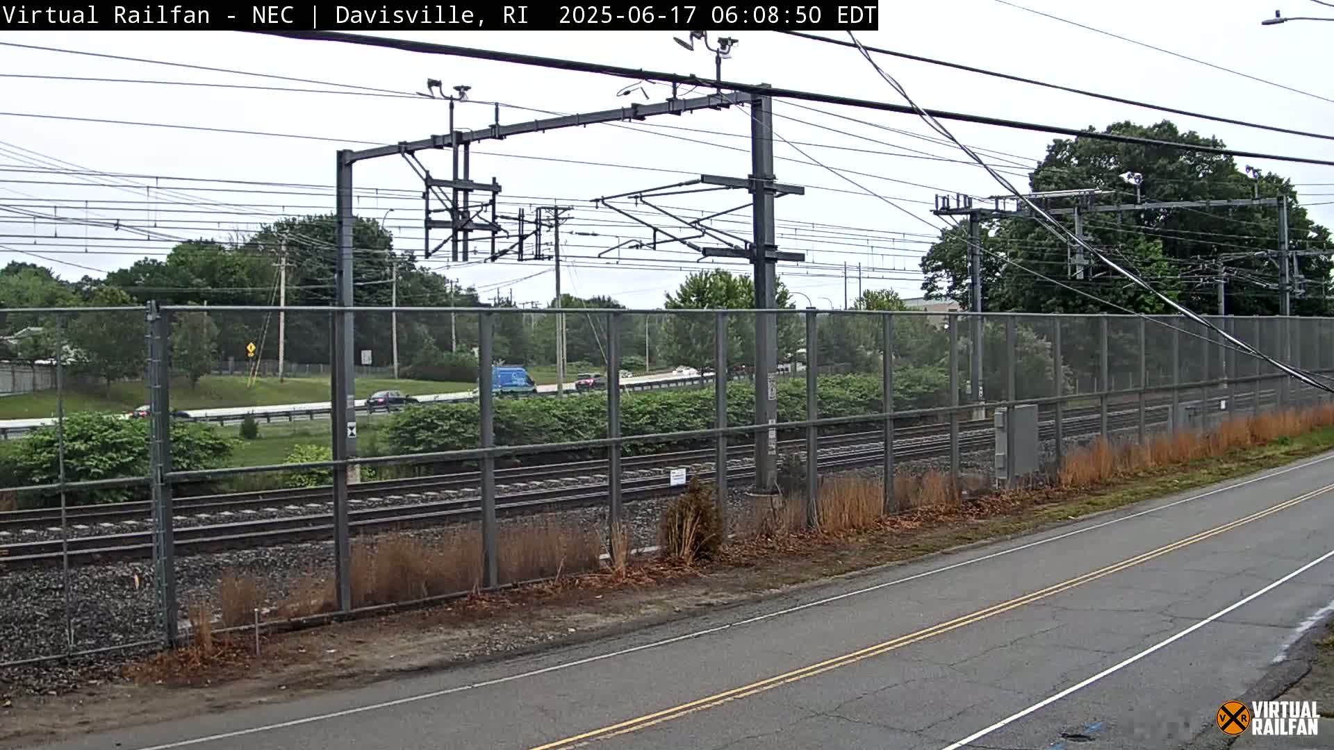 A view of railroad tracks and a road separated by a chain-link fence on an overcast day.