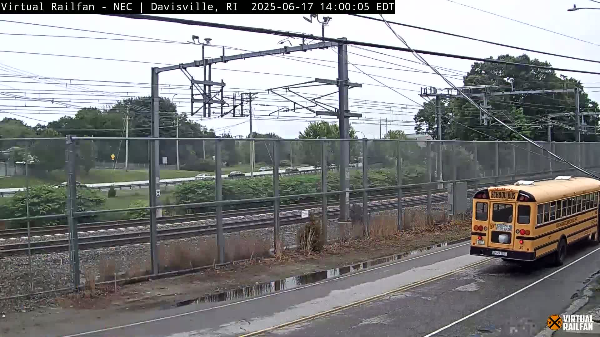 A yellow school bus drives on a road next to railroad tracks and a chain-link fence, under an overcast sky.