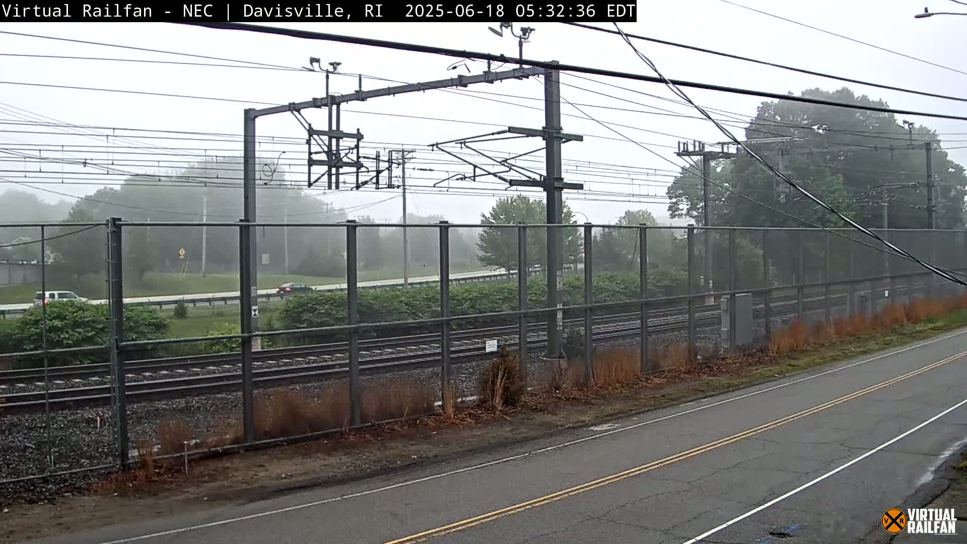 A misty view of railroad tracks alongside a road, separated by a chain-link fence, with trees and some vehicles visible in the background.