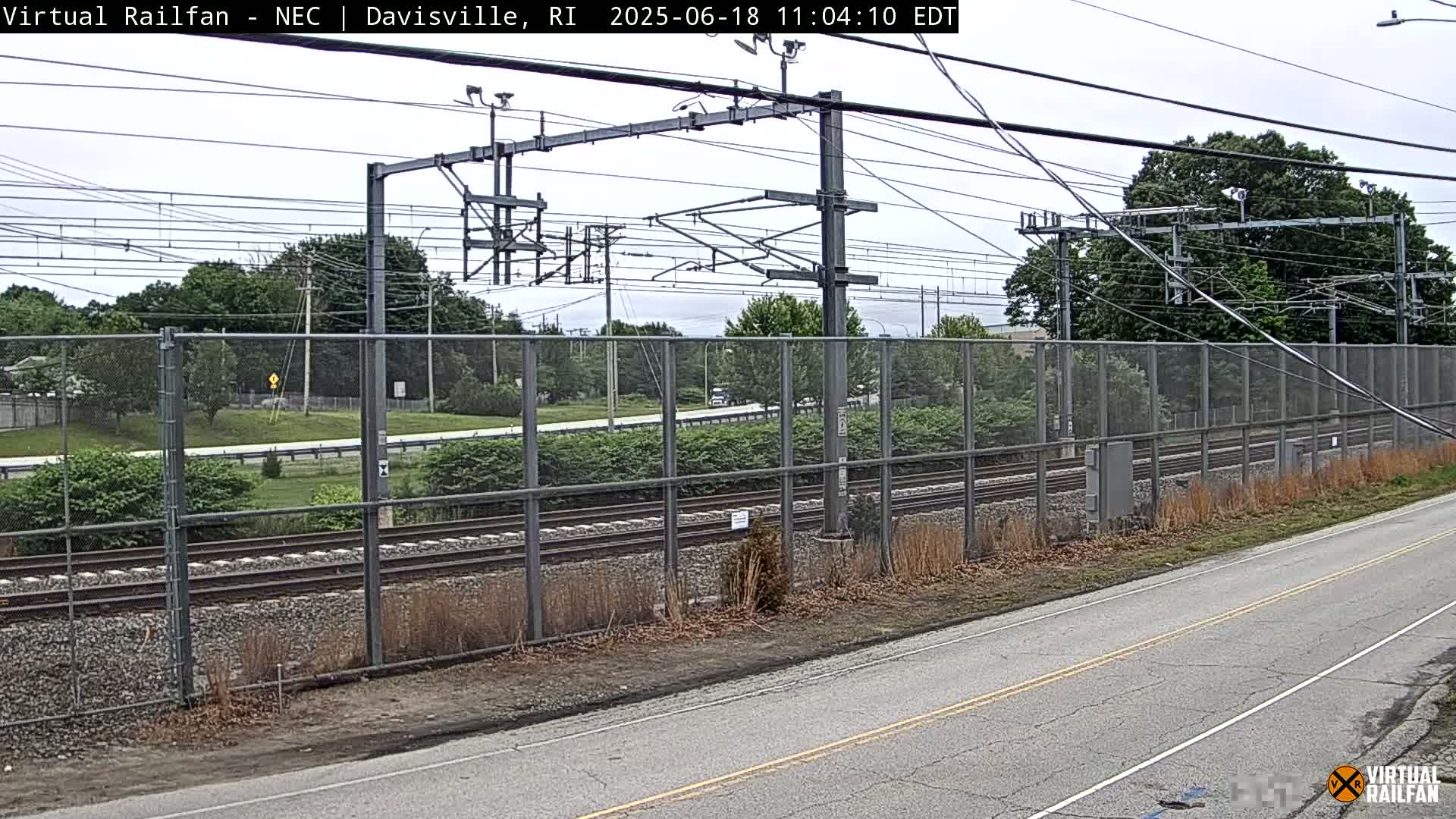 A section of train tracks runs alongside a road, separated by a chain-link fence, under a partly cloudy sky.