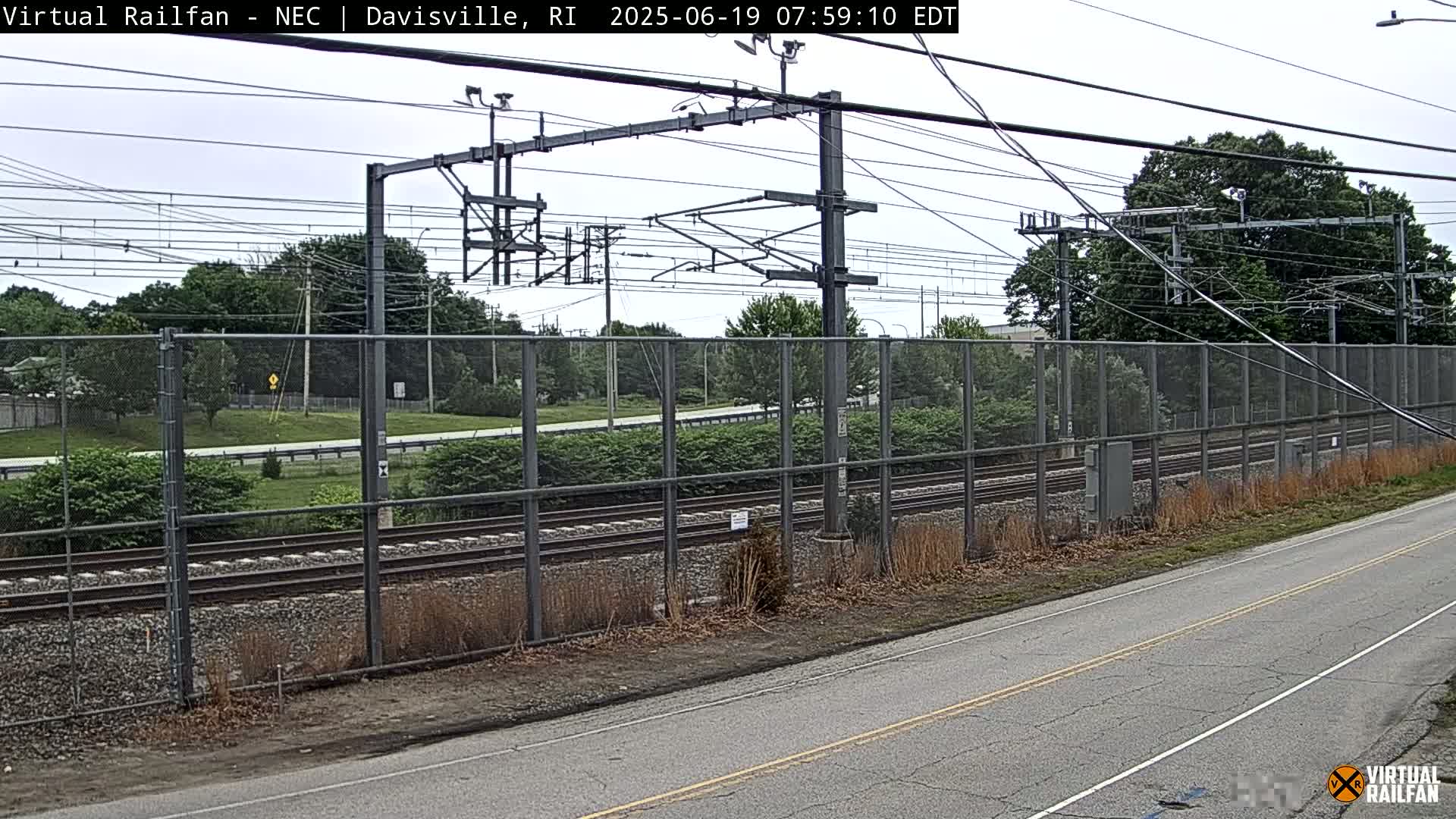 A view of railroad tracks behind a chain-link fence on an overcast day, with a road visible in the foreground.