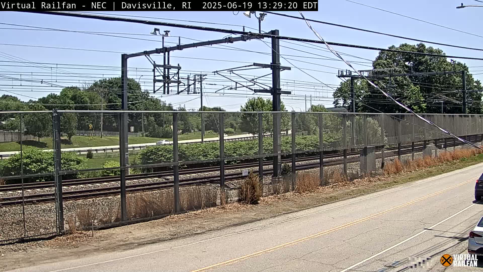 A sunny day view of train tracks behind a chain-link fence, beside a road with cars visible.
