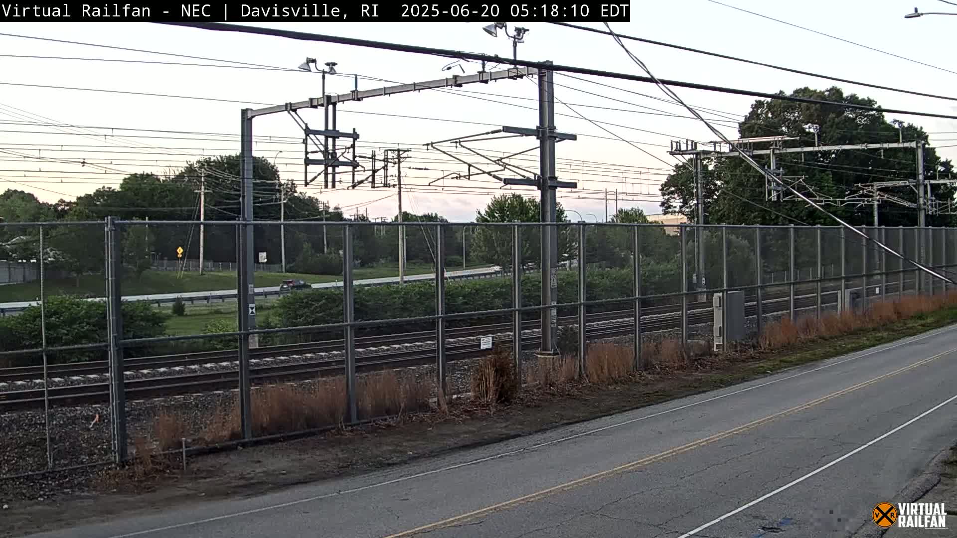 A view of railroad tracks behind a chain-link fence next to a road, under a partly cloudy sky.