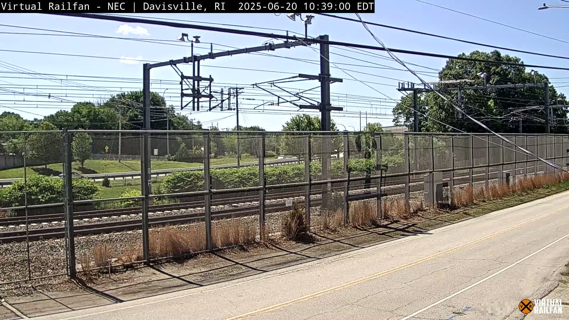 A sunny day reveals railroad tracks bordered by a chain-link fence, situated beside a road lined with vegetation.