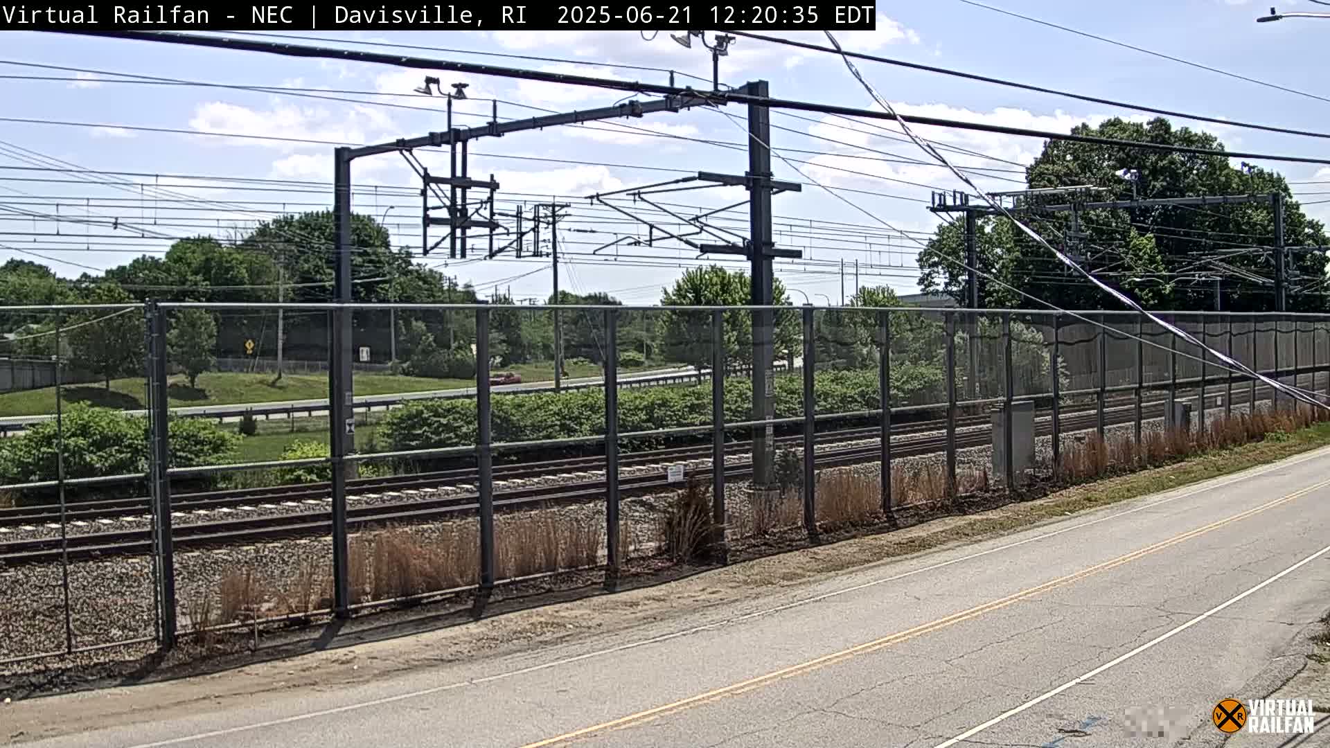 A view of railroad tracks behind a chain-link fence on a partly sunny day, with a road visible in the foreground.
