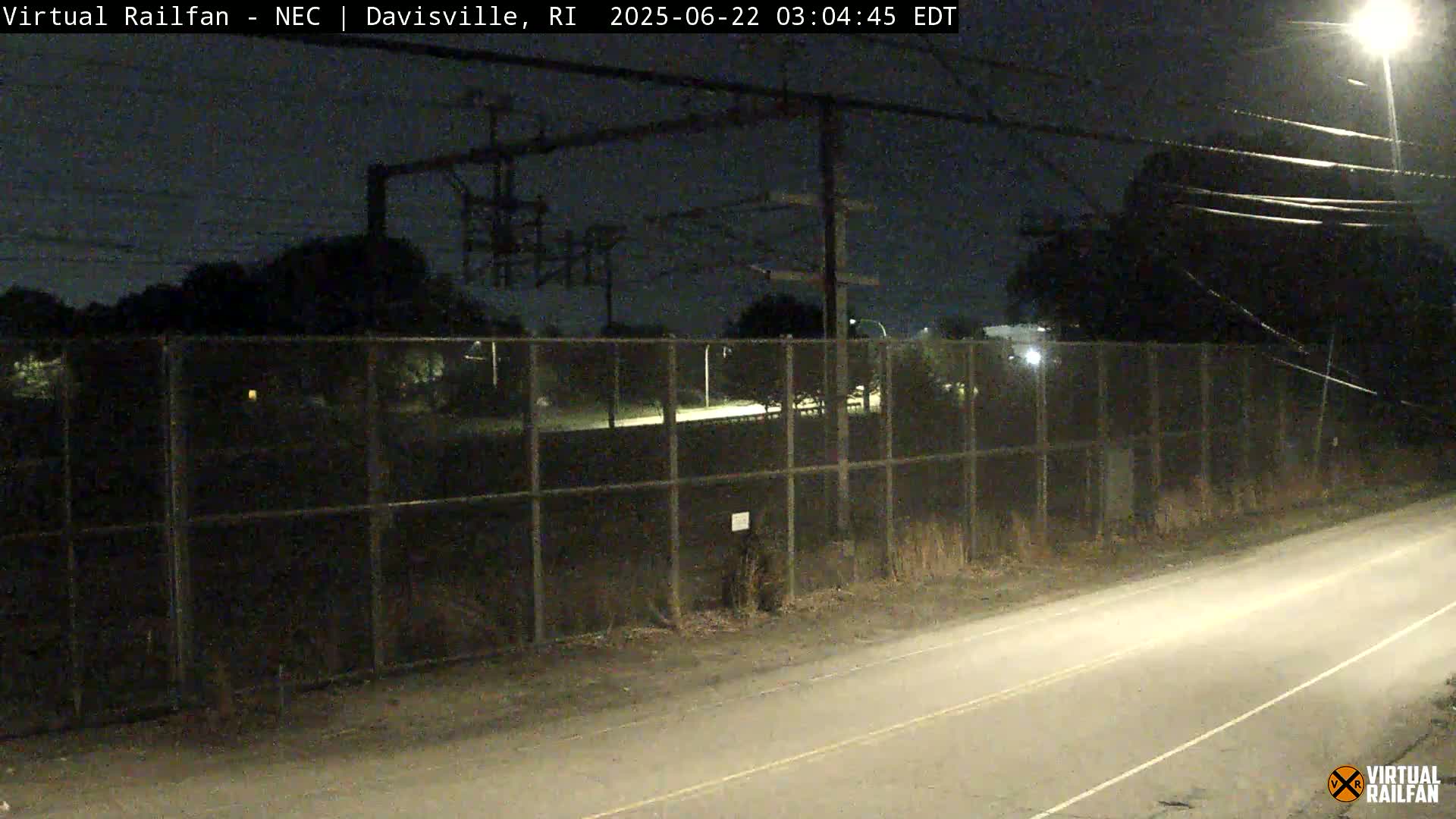 A nighttime view of a chain-link fence separating a road from a dark area with overhead power lines, under a mostly clear sky.