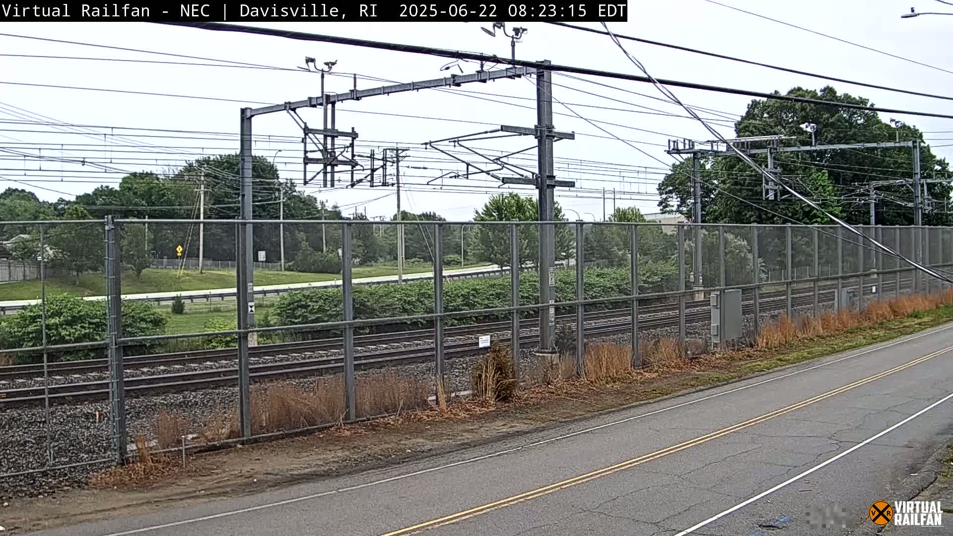 A view of railroad tracks behind a chain-link fence on an overcast day, with a road visible in the foreground.