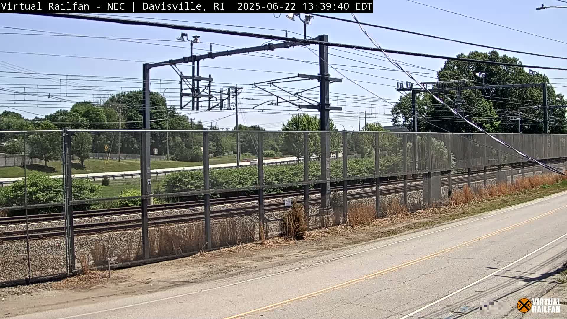 A view of train tracks behind a chain-link fence on a sunny day, with a road visible in the foreground.