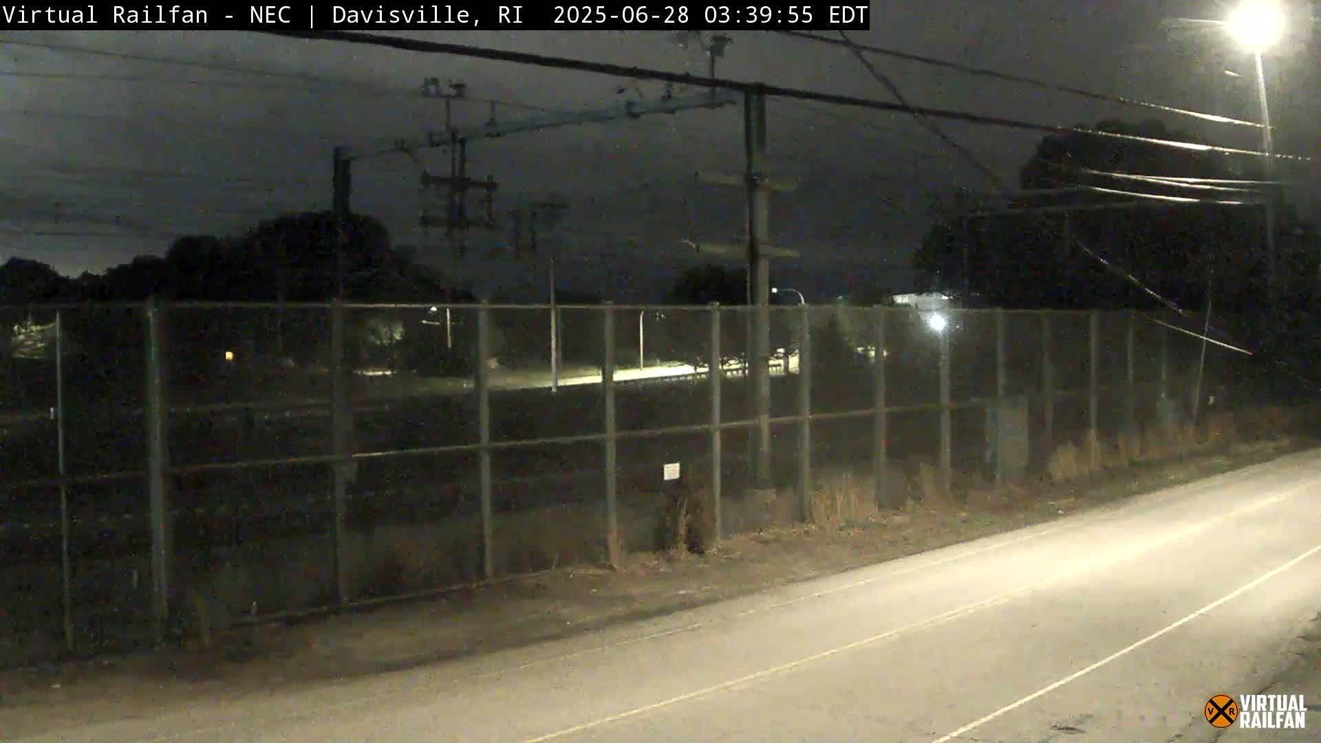 A nighttime view of a road next to a chain-link fence, beyond which are some trees and overhead power lines under a dark sky.