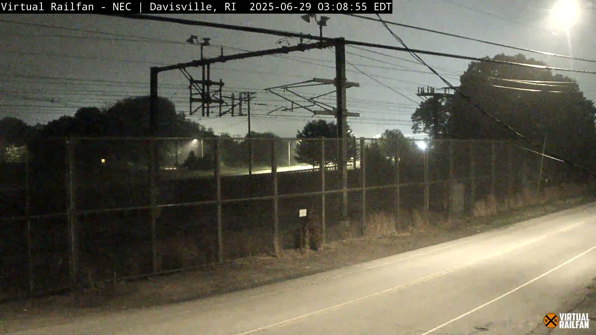 A nighttime view of a chain-link fence surrounding a dark area with power lines overhead, next to a road.