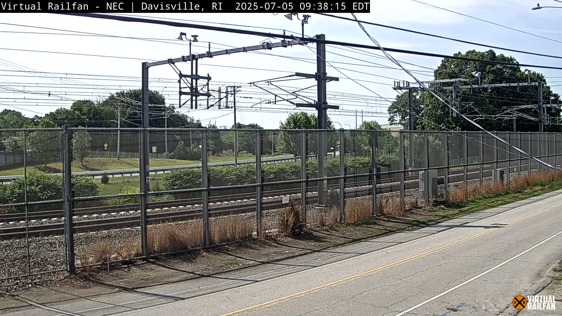 A view of railroad tracks behind a chain-link fence on a sunny day, with a road running parallel to the tracks in the foreground.