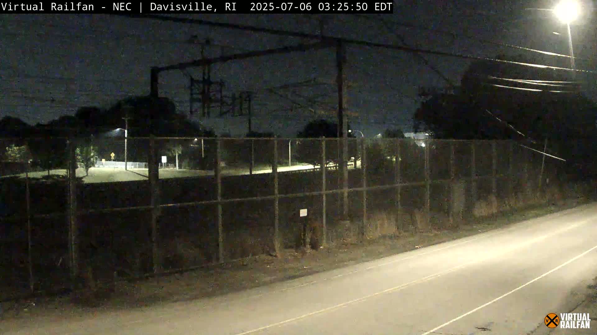 A nighttime view of a road next to a chain-link fence enclosing a dark, empty field, with overhead power lines and streetlights visible.