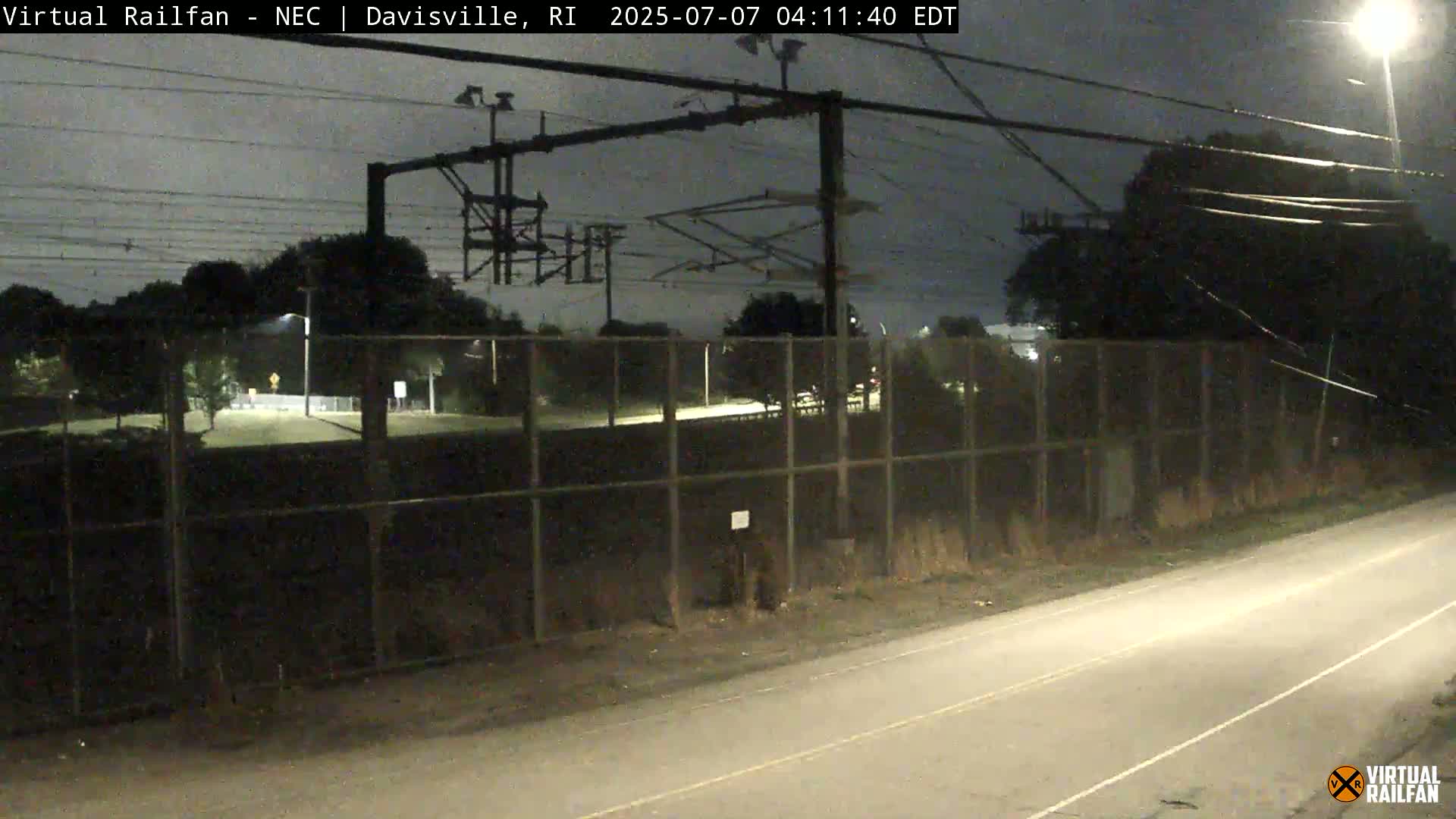 A nighttime view shows a chain-link fence separating a road from a dark, tree-lined area with overhead electrical wires and equipment.