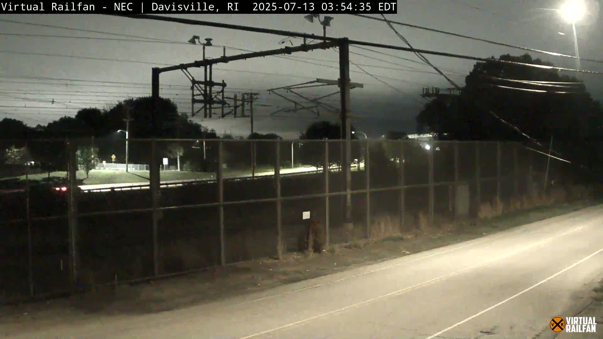 A nighttime view of a roadway next to a chain-link fence, beyond which are train tracks, electrical lines, and trees under a cloudy sky.