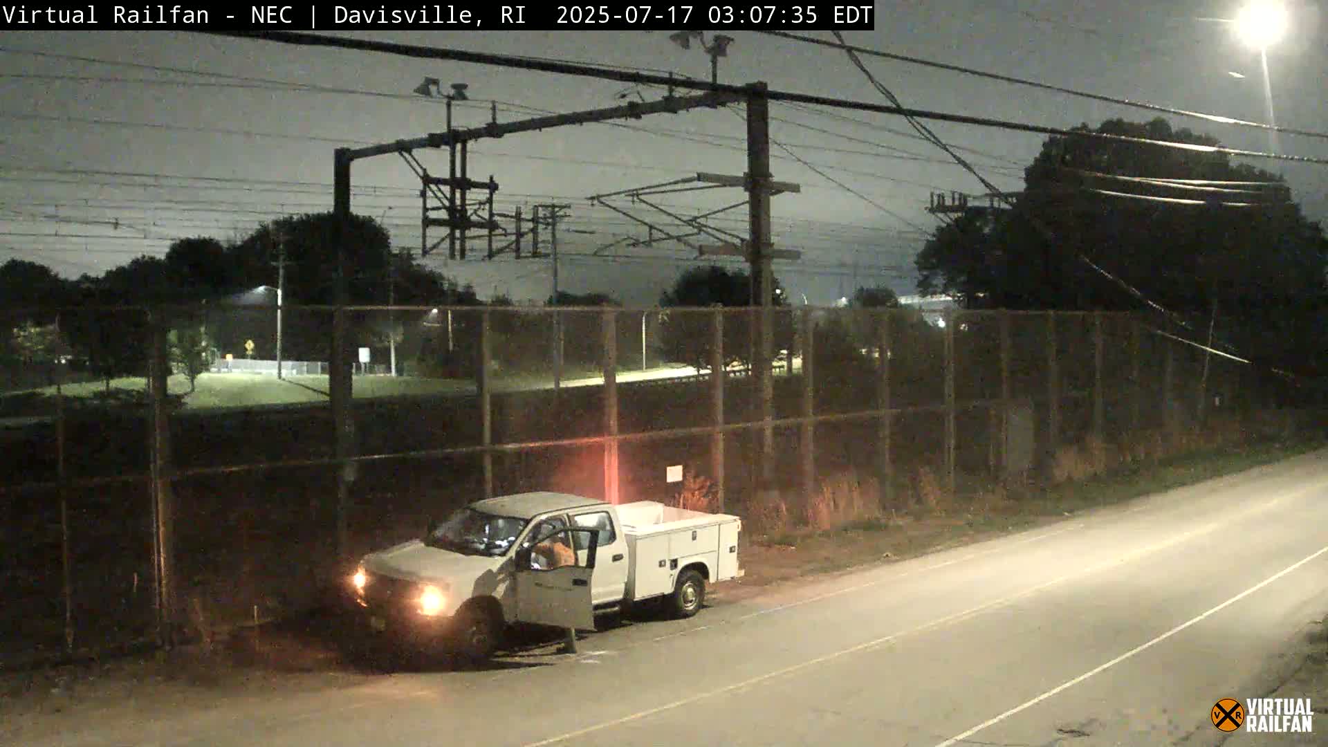 A white pickup truck with its door open is parked on a road next to a chain-link fence under a dark, overcast night sky.