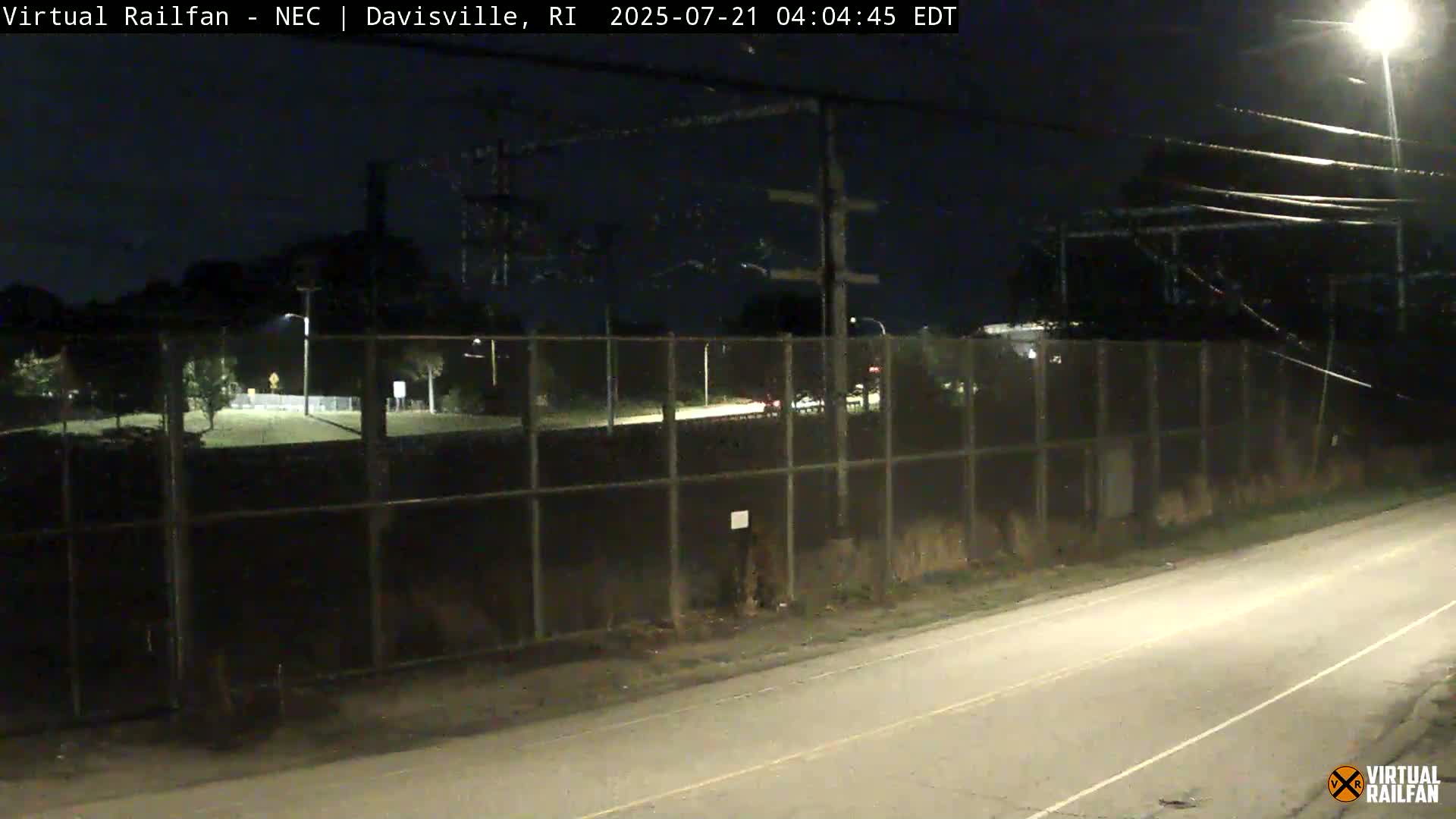 A nighttime view of a road next to a chain-link fence enclosing a dark, empty field, with a few cars visible in the distance.