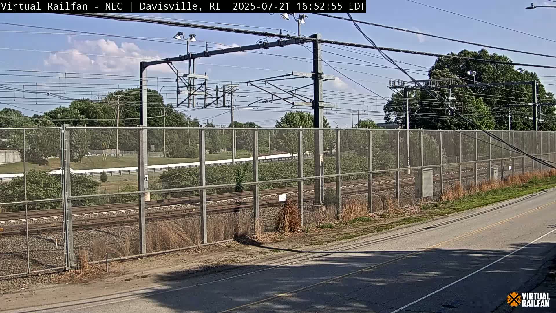 A view of railroad tracks behind a chain-link fence on a sunny day with partly cloudy skies, next to a road.