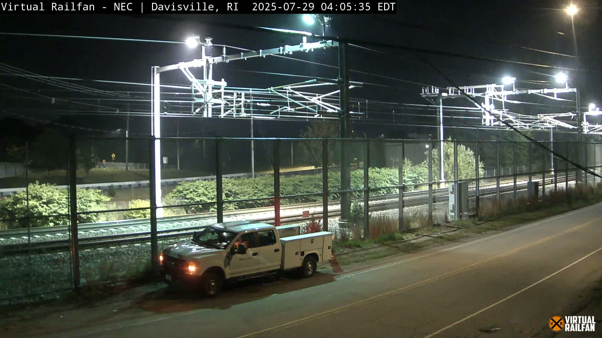 A white pickup truck is parked next to a chain-link fence alongside a set of train tracks at night under bright lights.