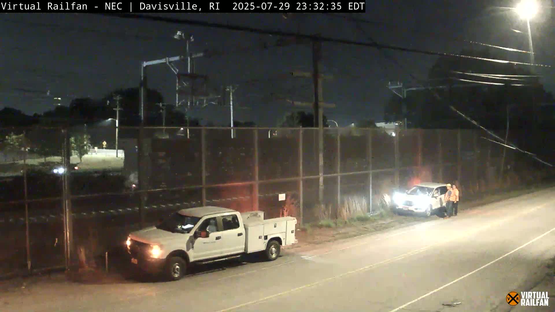 A white pickup truck is parked next to a chain-link fence alongside a set of train tracks at night under bright lights.
