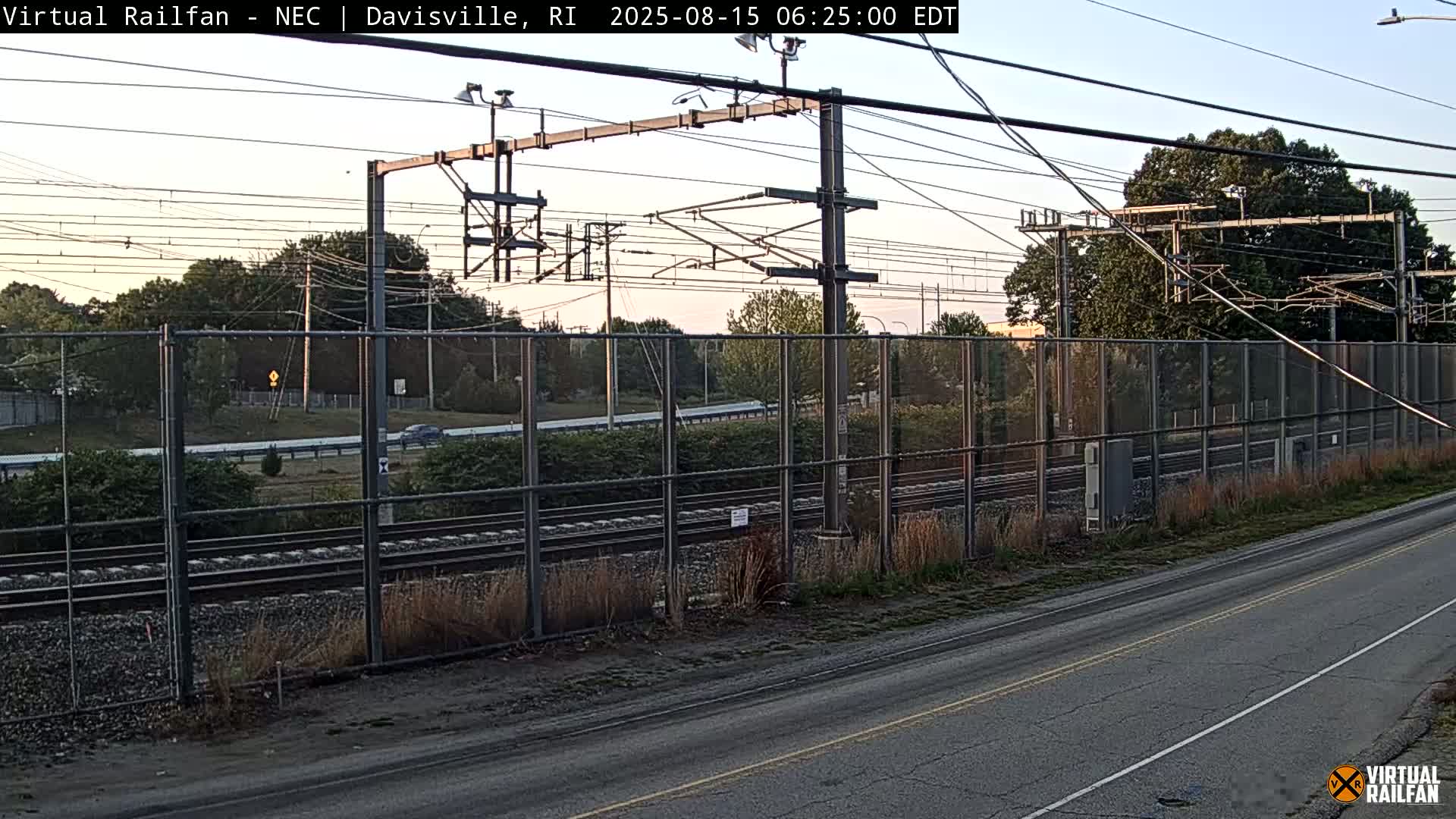 A white pickup truck is parked next to a chain-link fence alongside a set of train tracks at night under bright lights.