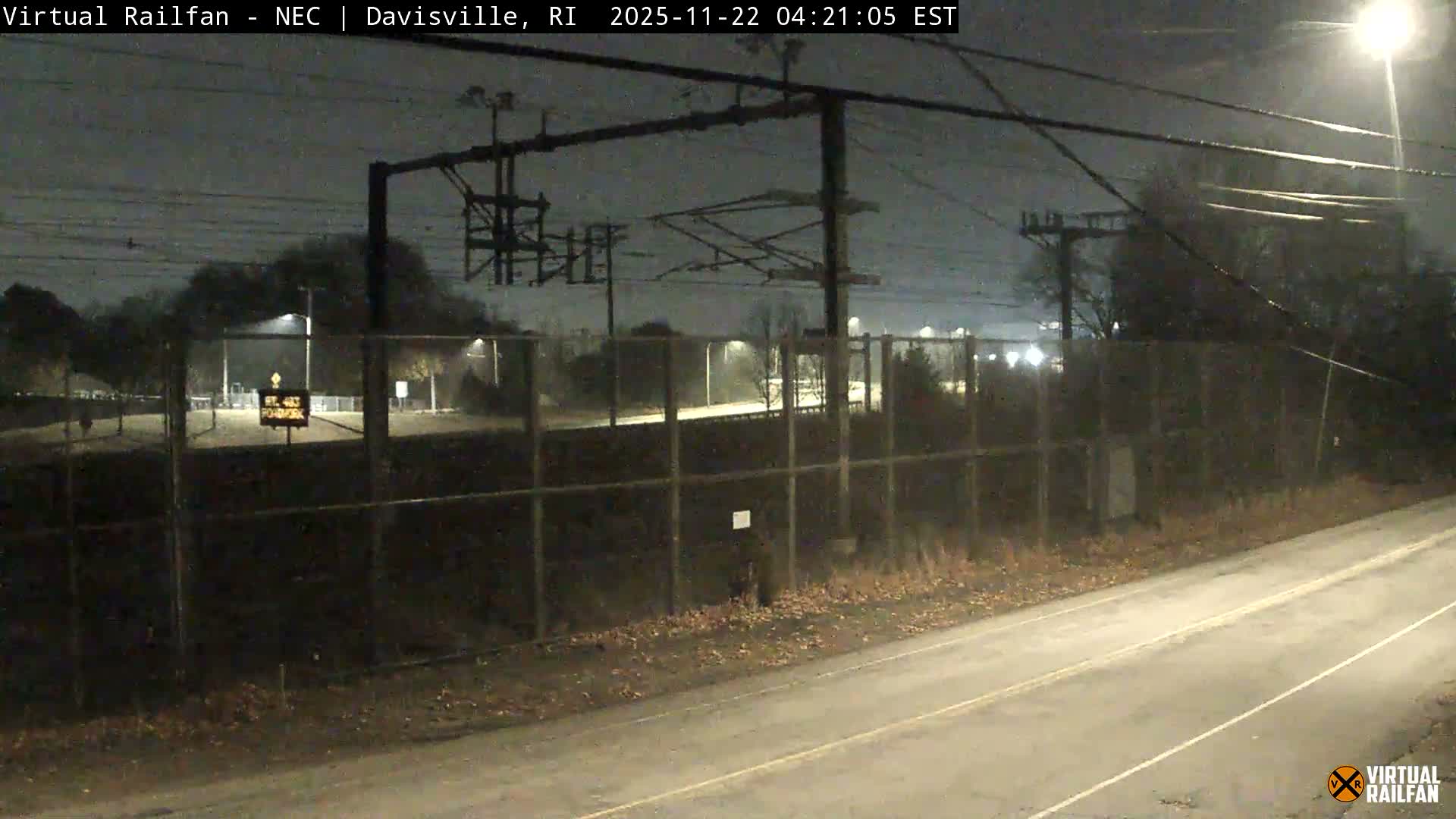 An overcast and damp early morning scene displays a paved road in the foreground, a chain-link fence running across the middle ground, and complex railway power line structures overhead, with distant lights illuminating trees and fields beyond.