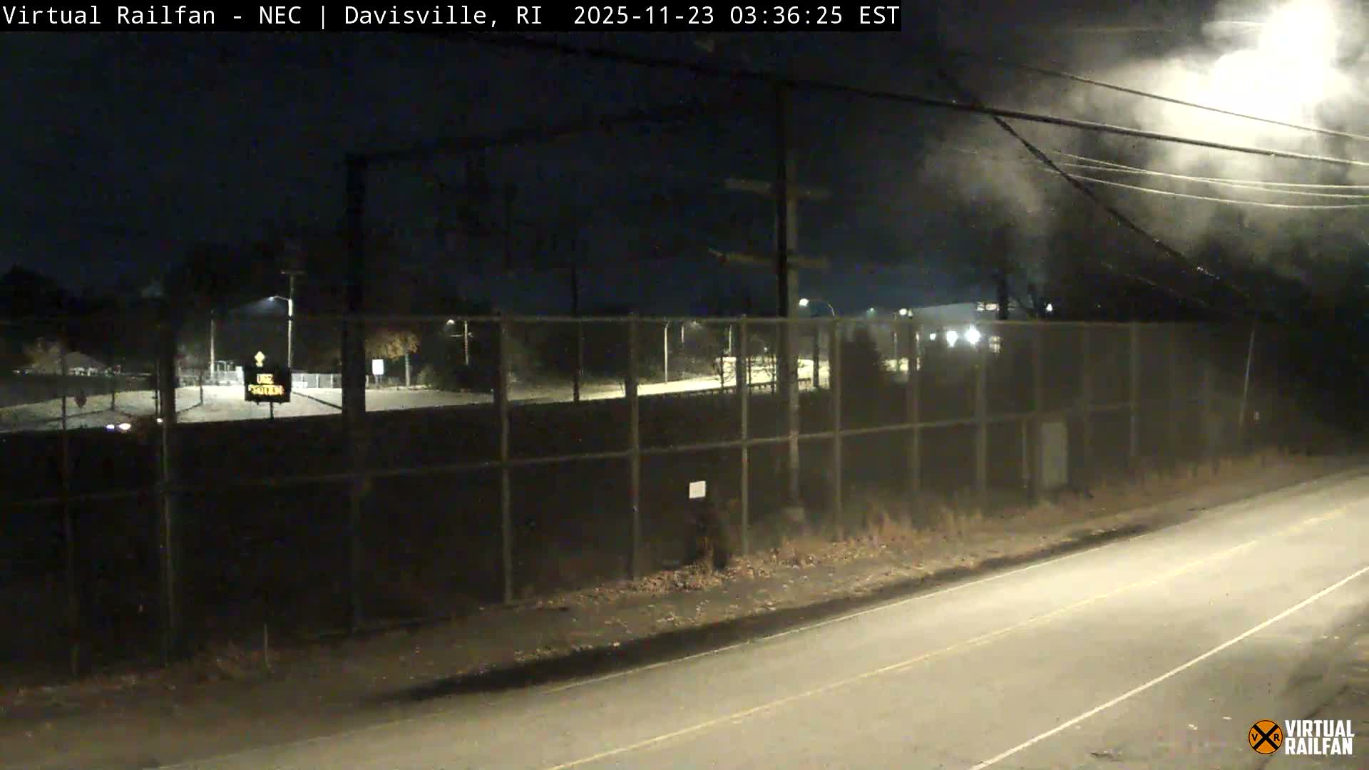 A dark night scene reveals a paved road in the foreground, a long chain-link fence, and distant lights illuminating trees and structures, with notable mist or fog present, especially in the upper right.