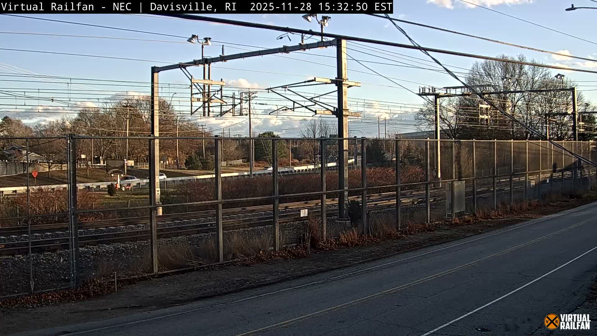 A rail line with overhead catenary wires and a chain-link fence runs alongside an empty road in the foreground, under a clear, sunny sky with scattered white clouds.
