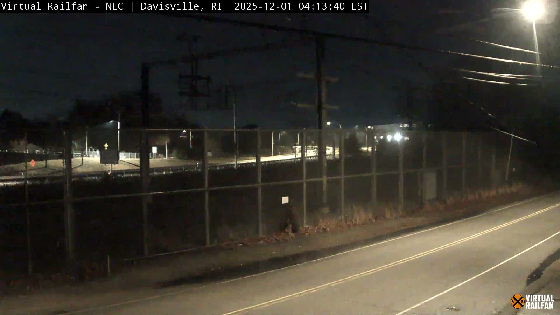 A clear night scene shows a streetlight-illuminated paved road next to a chain-link fence, beyond which lies a dimly visible industrial or transport area with utility poles and distant lights.