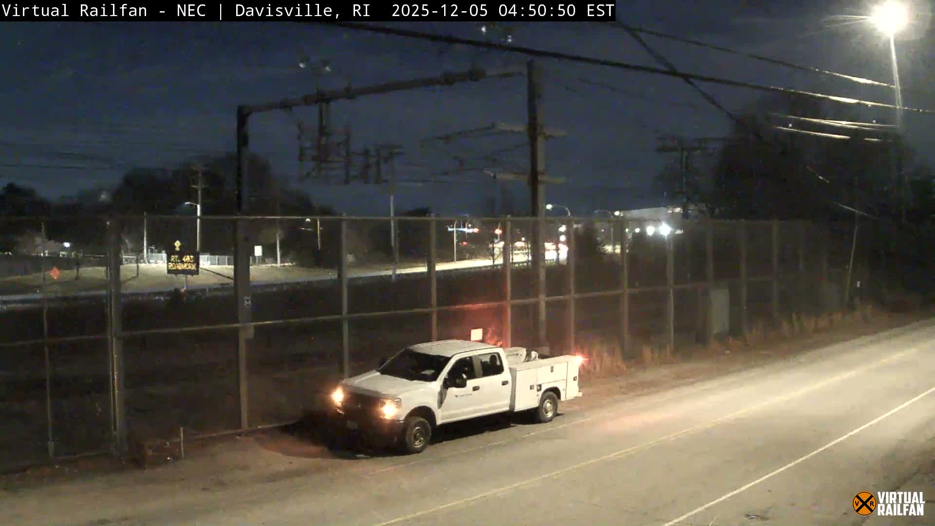 A white utility truck with its hazard lights illuminated is stopped on a dirt shoulder next to a road and a chain-link fence under a clear night sky, with streetlights and distant highway lights visible in the background.