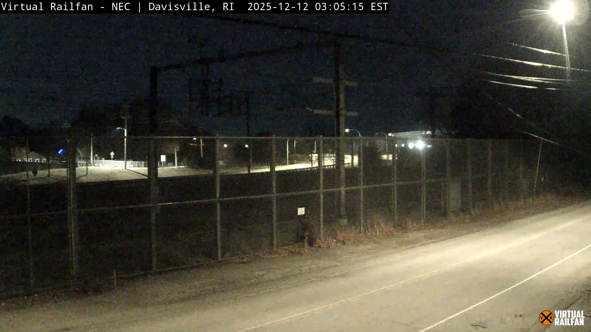 A clear night reveals an outdoor scene with a paved road in the foreground, bordered by a chain-link fence, and distant structures marked by scattered lights and utility poles.