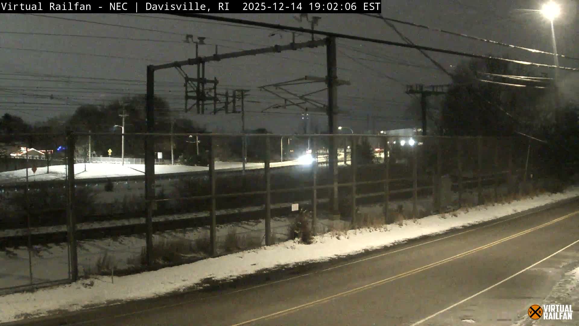 A clear night reveals an outdoor scene with a paved road in the foreground, bordered by a chain-link fence, and distant structures marked by scattered lights and utility poles.