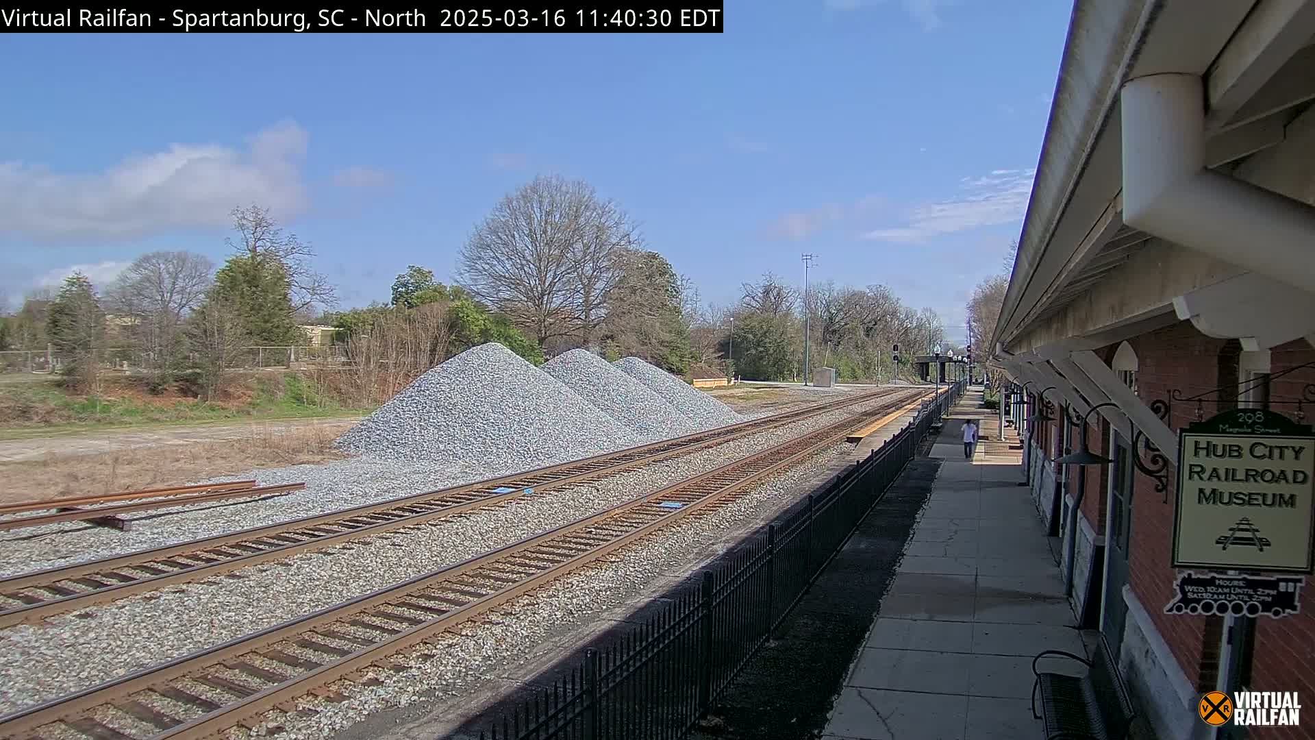 A sunny day reveals railroad tracks with piles of gravel beside them, alongside a brick railroad museum.