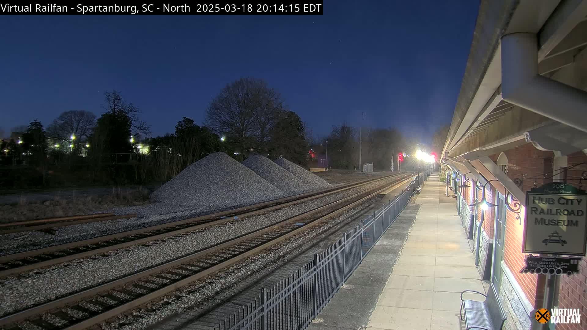 Under a dark twilight sky, railroad tracks extend from a brick train station toward piles of gravel.