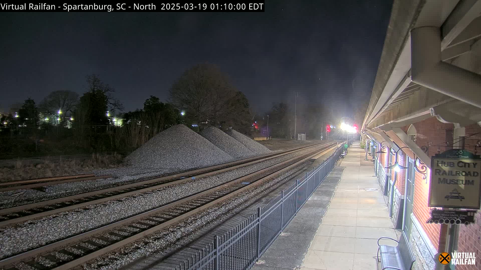 Under a dark night sky, train tracks extend from a nighttime train station platform with piles of gravel nearby.