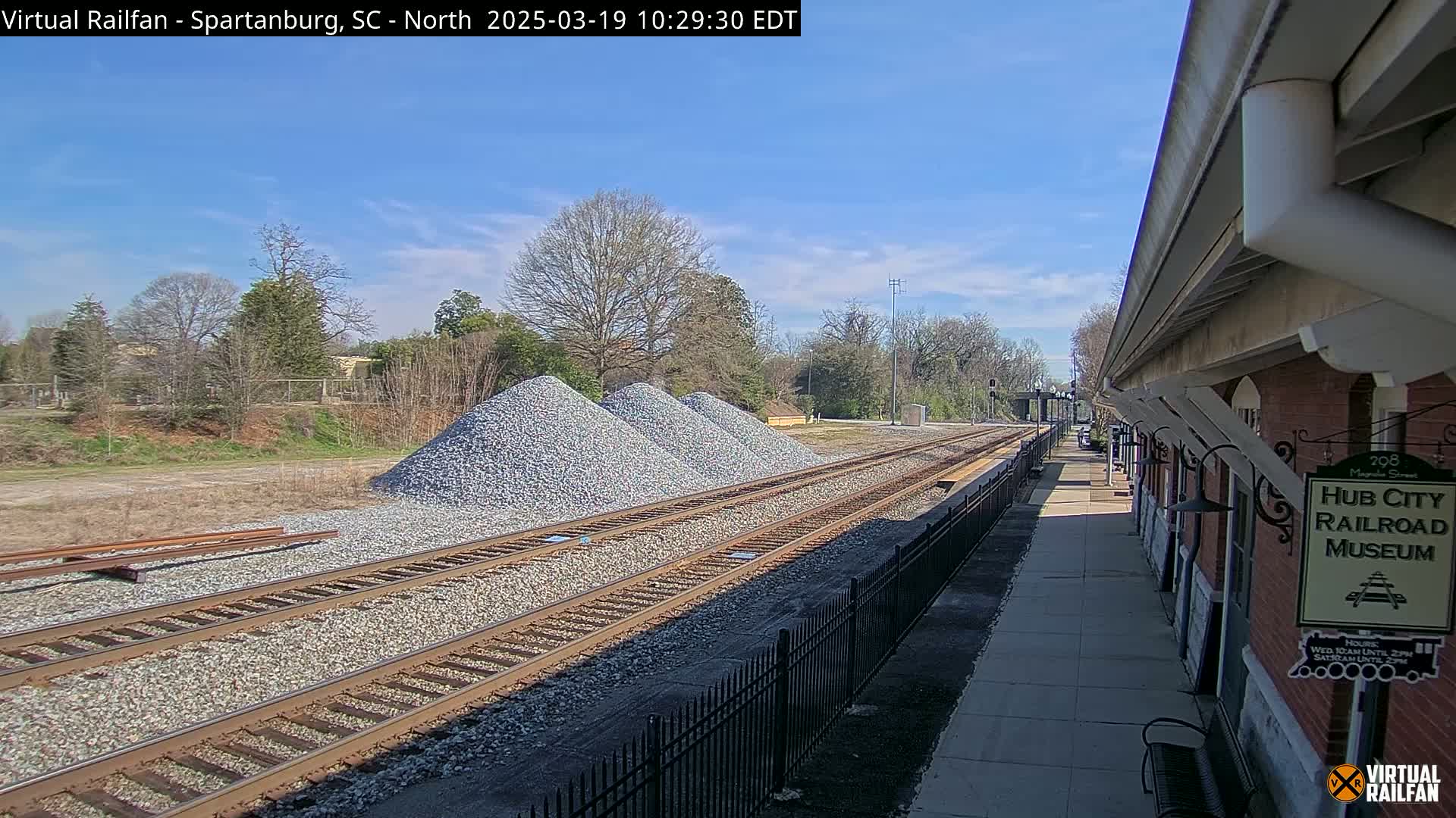 Under a mostly sunny sky, railroad tracks run alongside a train platform next to piles of gravel.