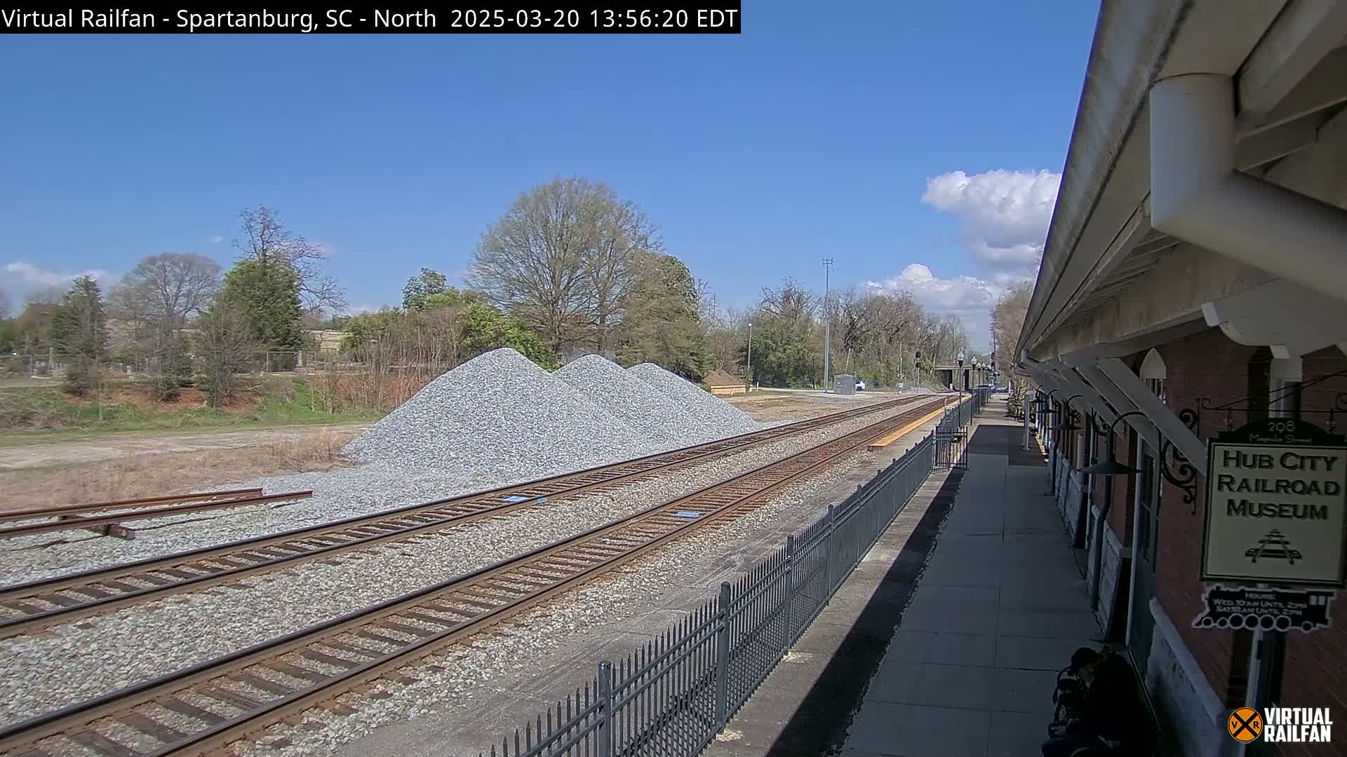 A sunny day reveals railroad tracks next to a platform, with piles of gravel nearby and trees in the background.