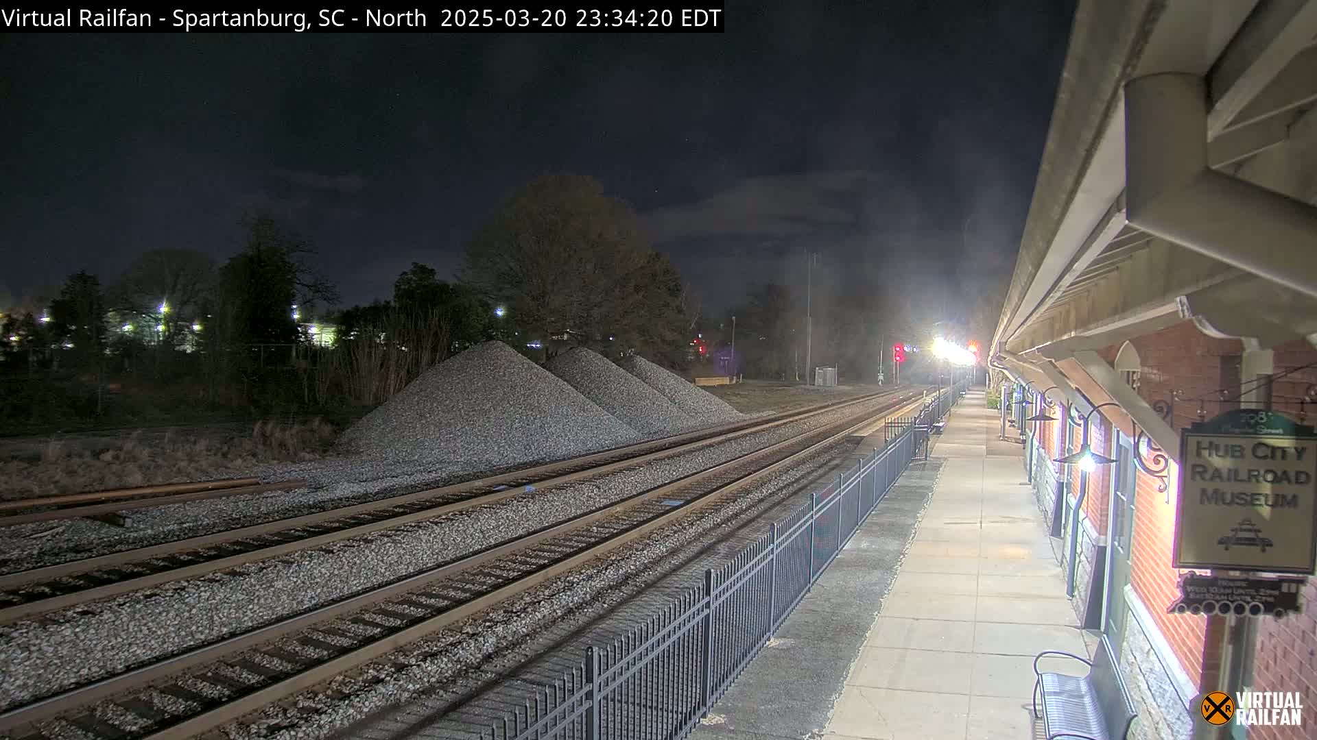 At night, a train station platform extends alongside railroad tracks leading to piles of gravel under a dark, clear sky.